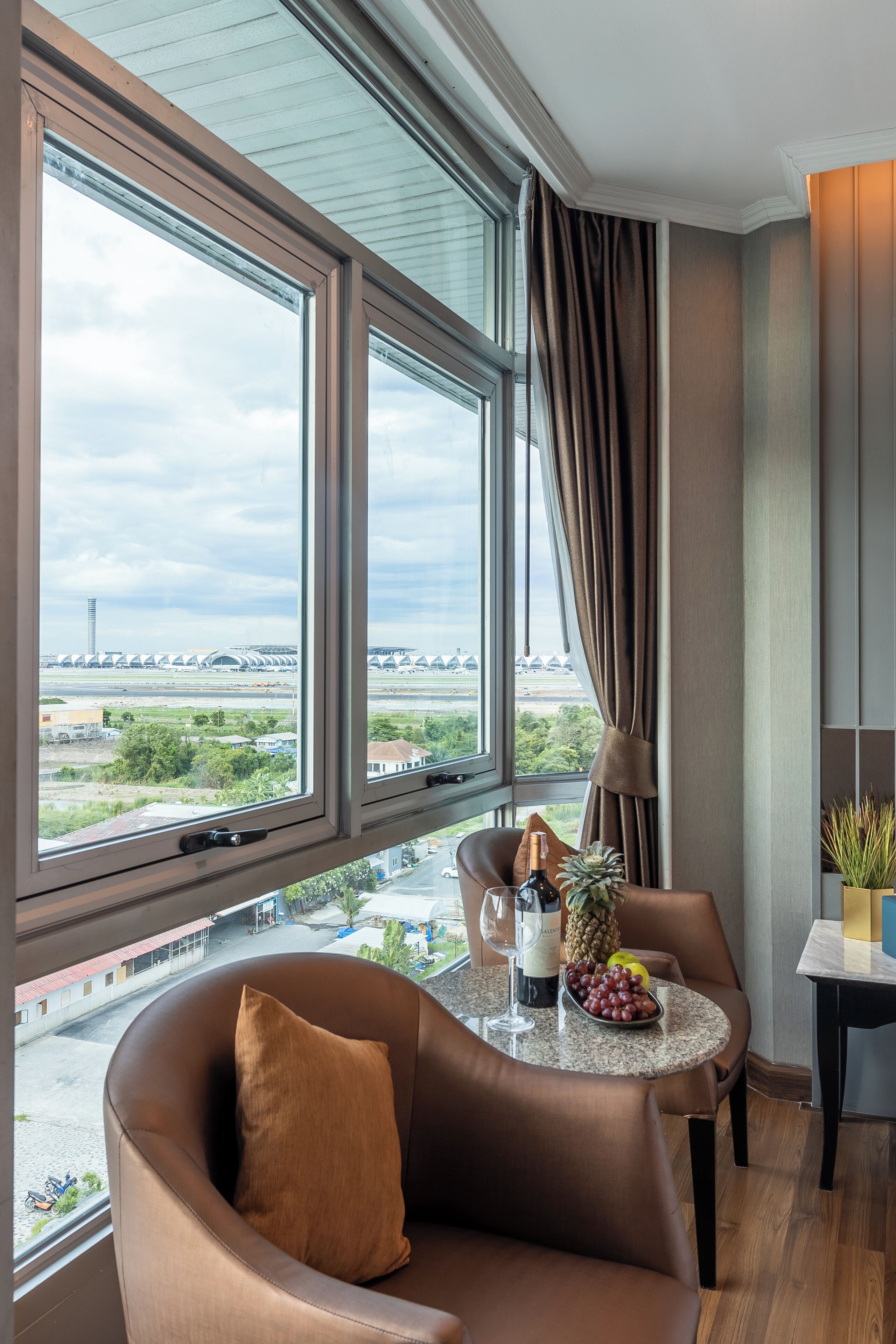 Una habitación de hotel moderna con ventanas amplias, un sillón de cuero confortable, una mesita redonda con una botella de vino, un cuenco de fruta y una planta en maceta.