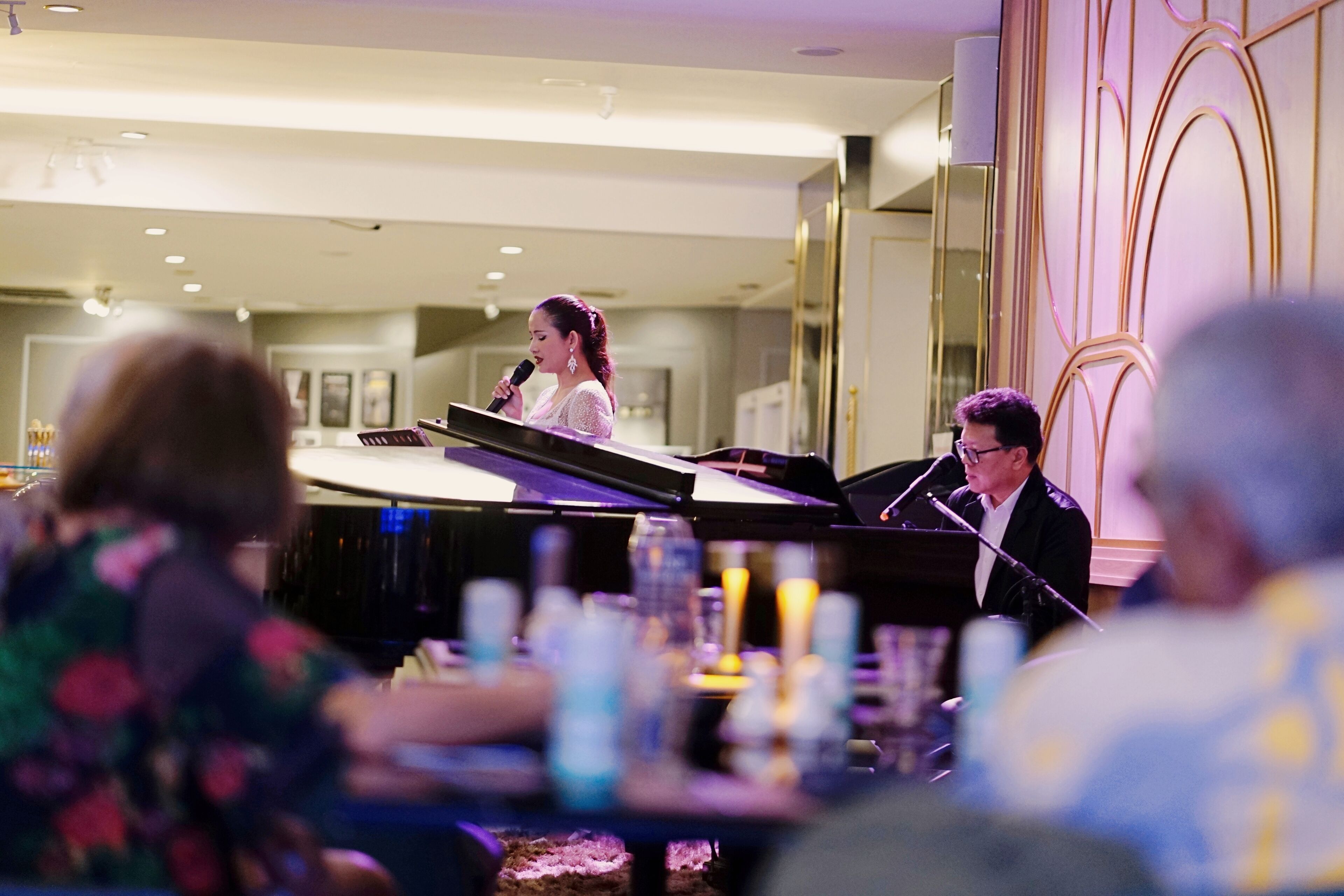 A woman speaking at a podium in a hotel lobby with a piano and a man seated at the piano.