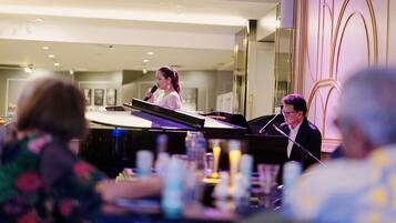 A woman speaking at a podium in a hotel lobby with a piano and a man seated at the piano.