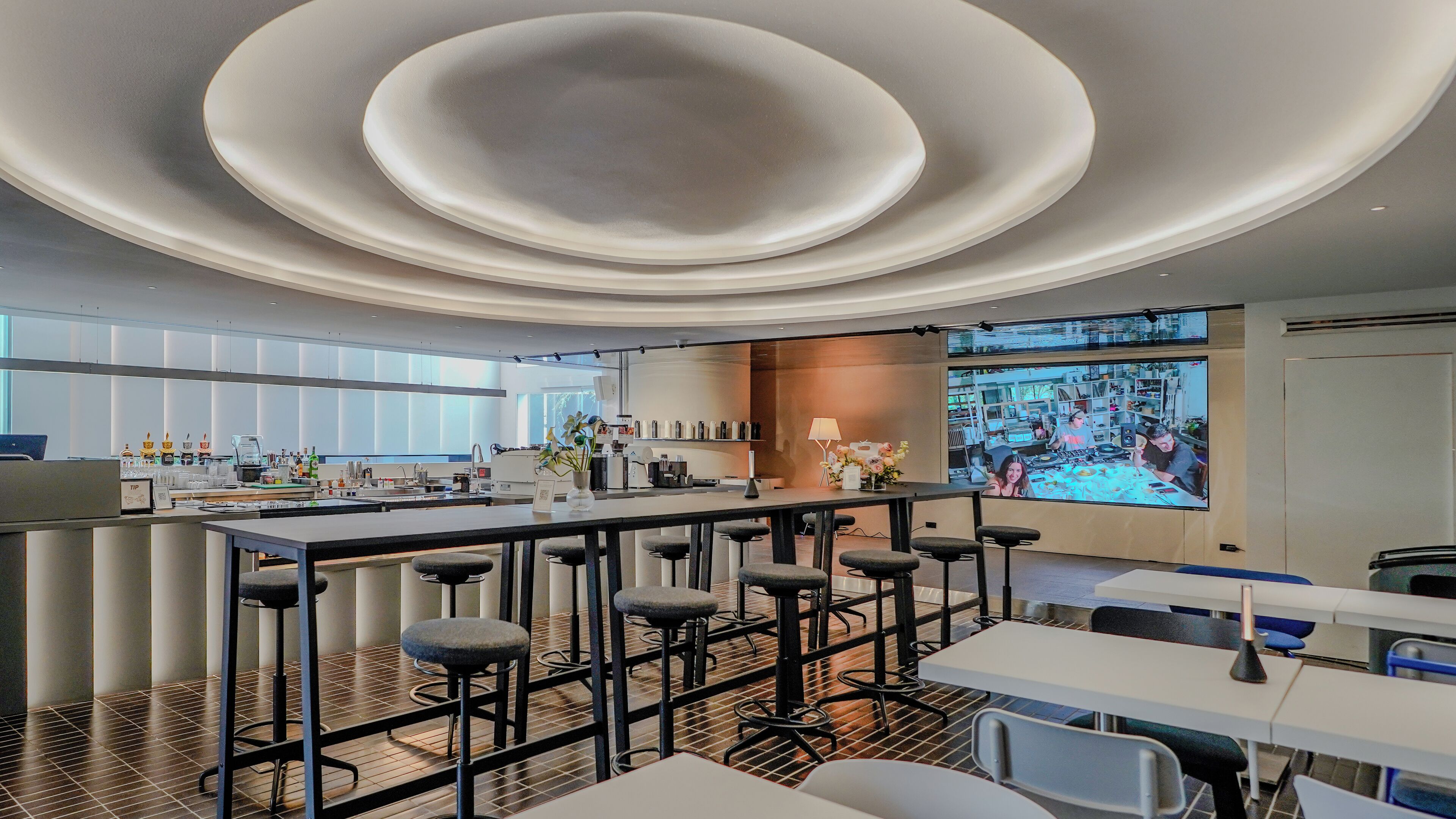 Zona de bar moderna con barra central, varios taburetes y una pantalla grande mostrando un evento deportivo.