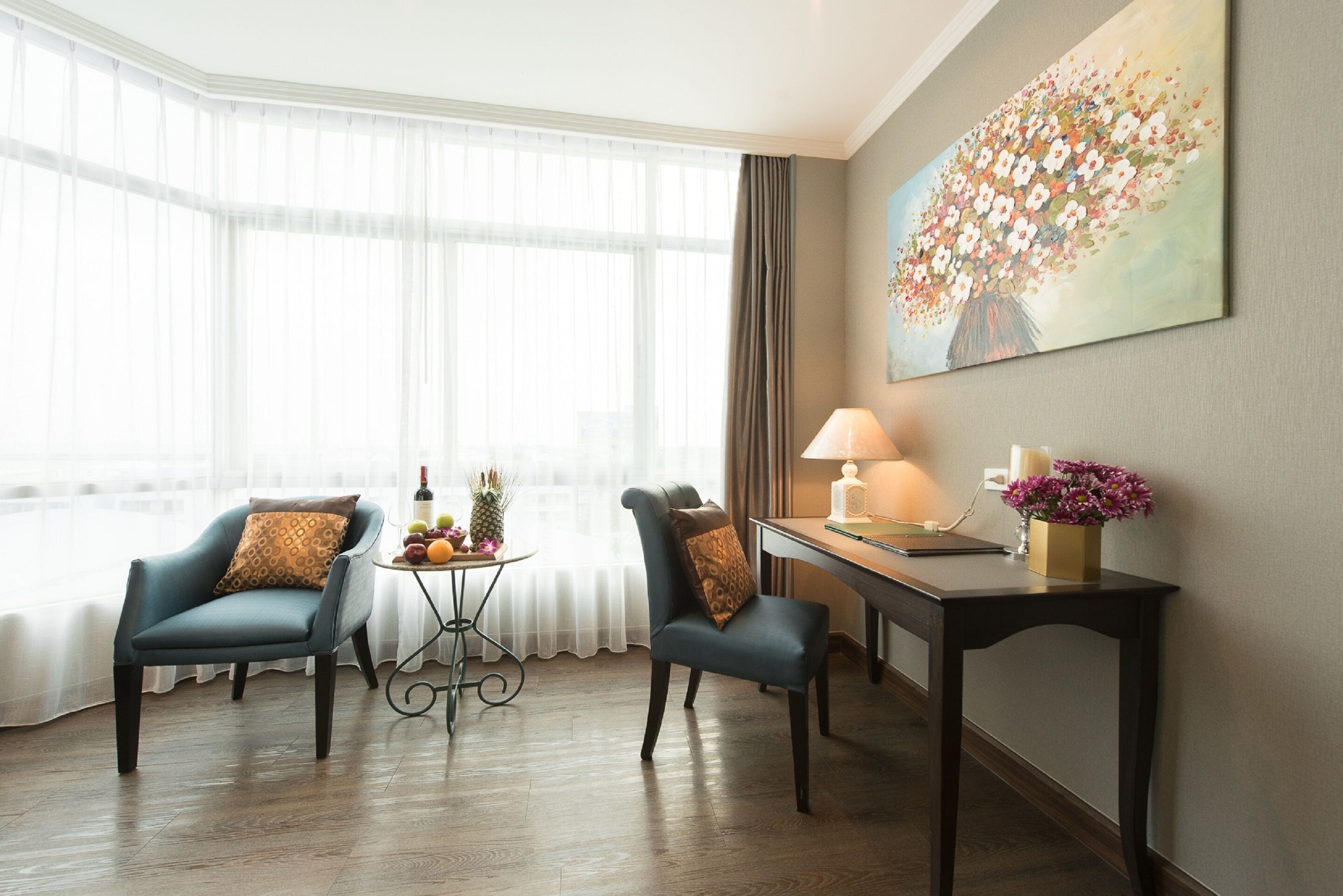 Une chambre d’hôtel avec deux chaises, une petite table sur laquelle se trouvent des fruits et une bouteille, un bureau avec une lampe et un tableau accroché au mur.