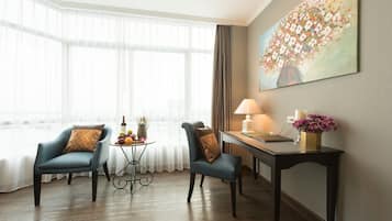 Une chambre d’hôtel avec deux chaises, une petite table sur laquelle se trouvent des fruits et une bouteille, un bureau avec une lampe et un tableau accroché au mur.