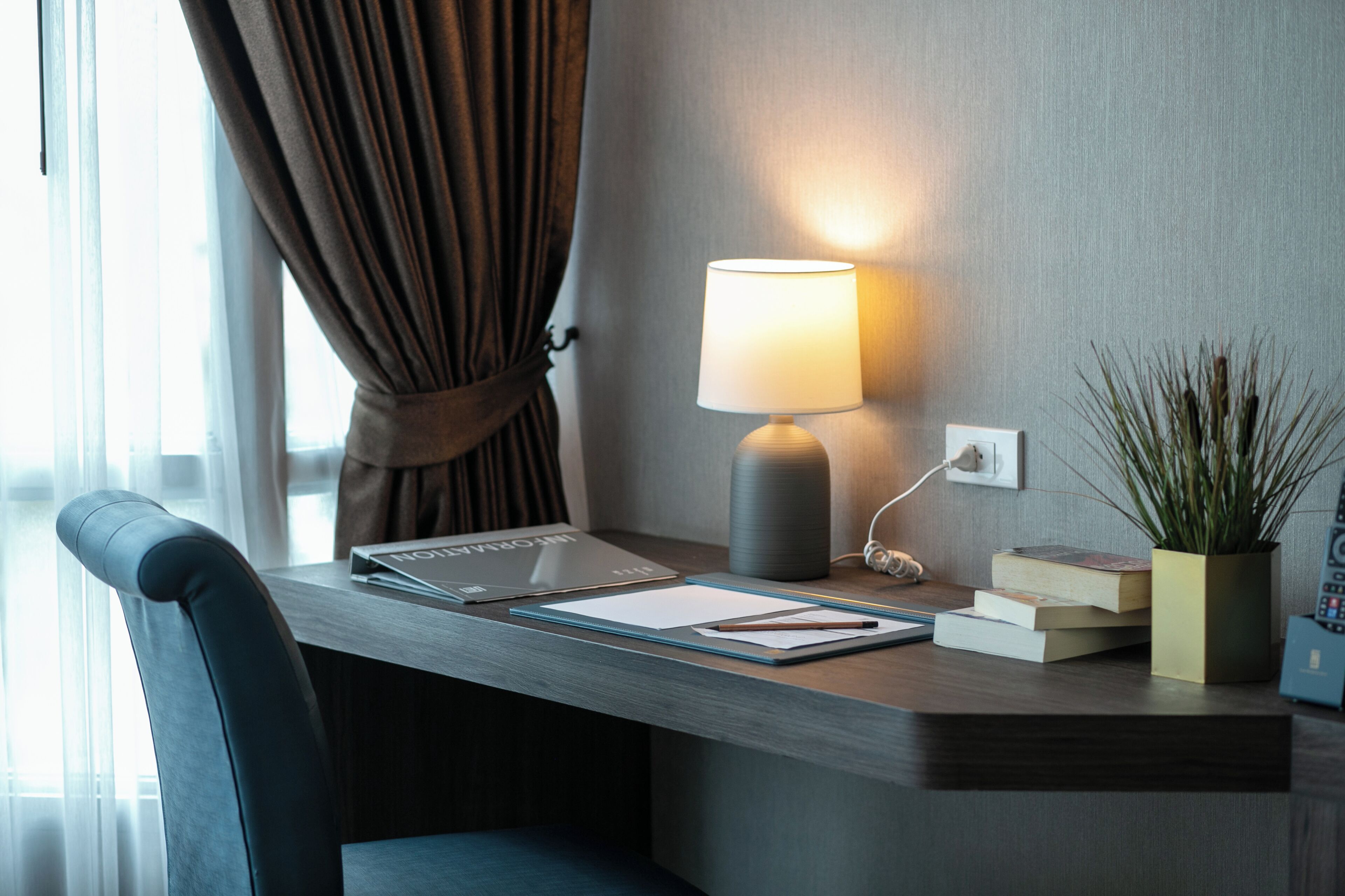 Ein Hotel-Schreibtisch mit Lampe, Laptop, Büchern und einer Topfpflanze.