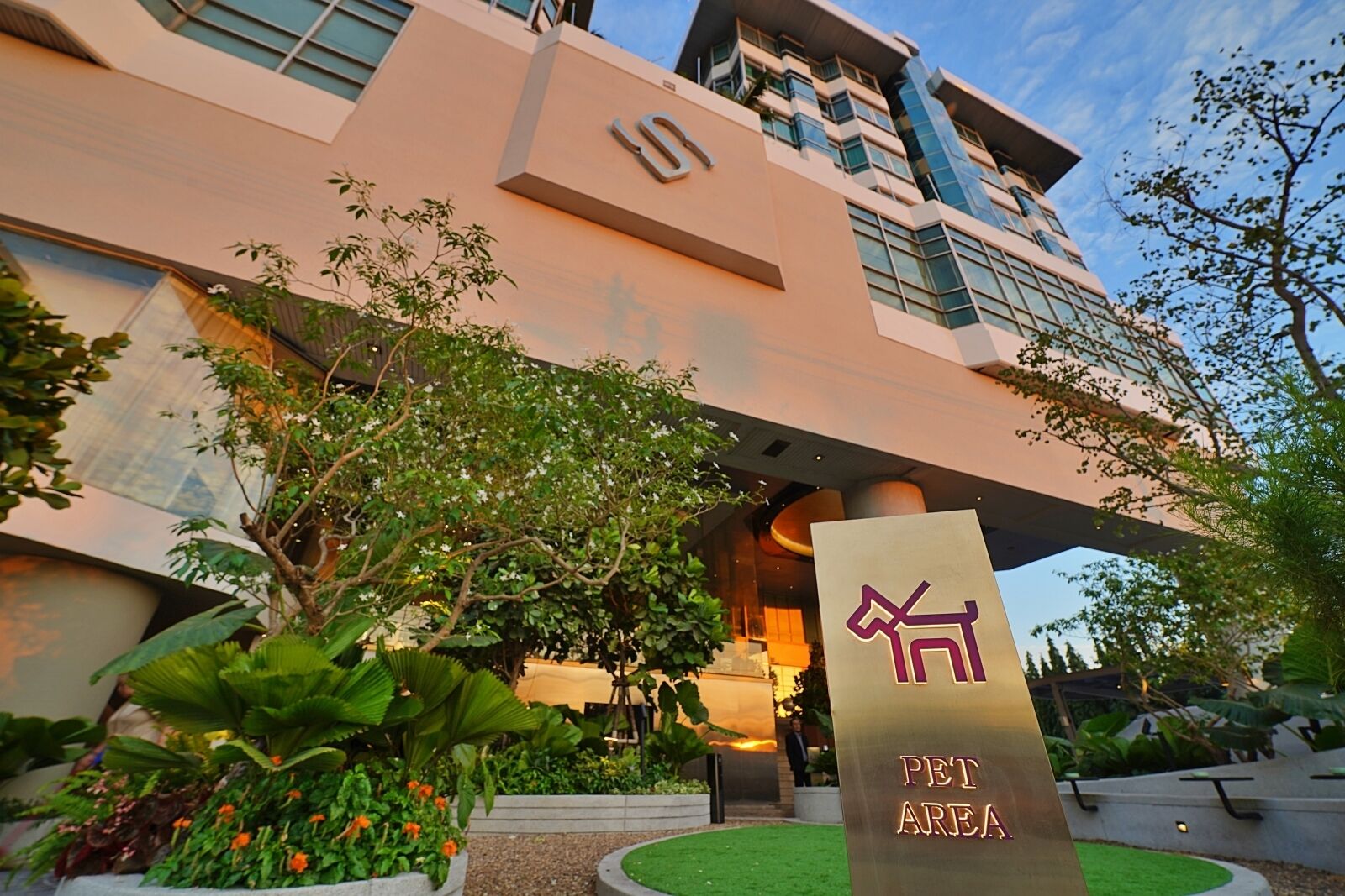 A modern building with a pet area sign, surrounded by lush greenery and a well-maintained garden.