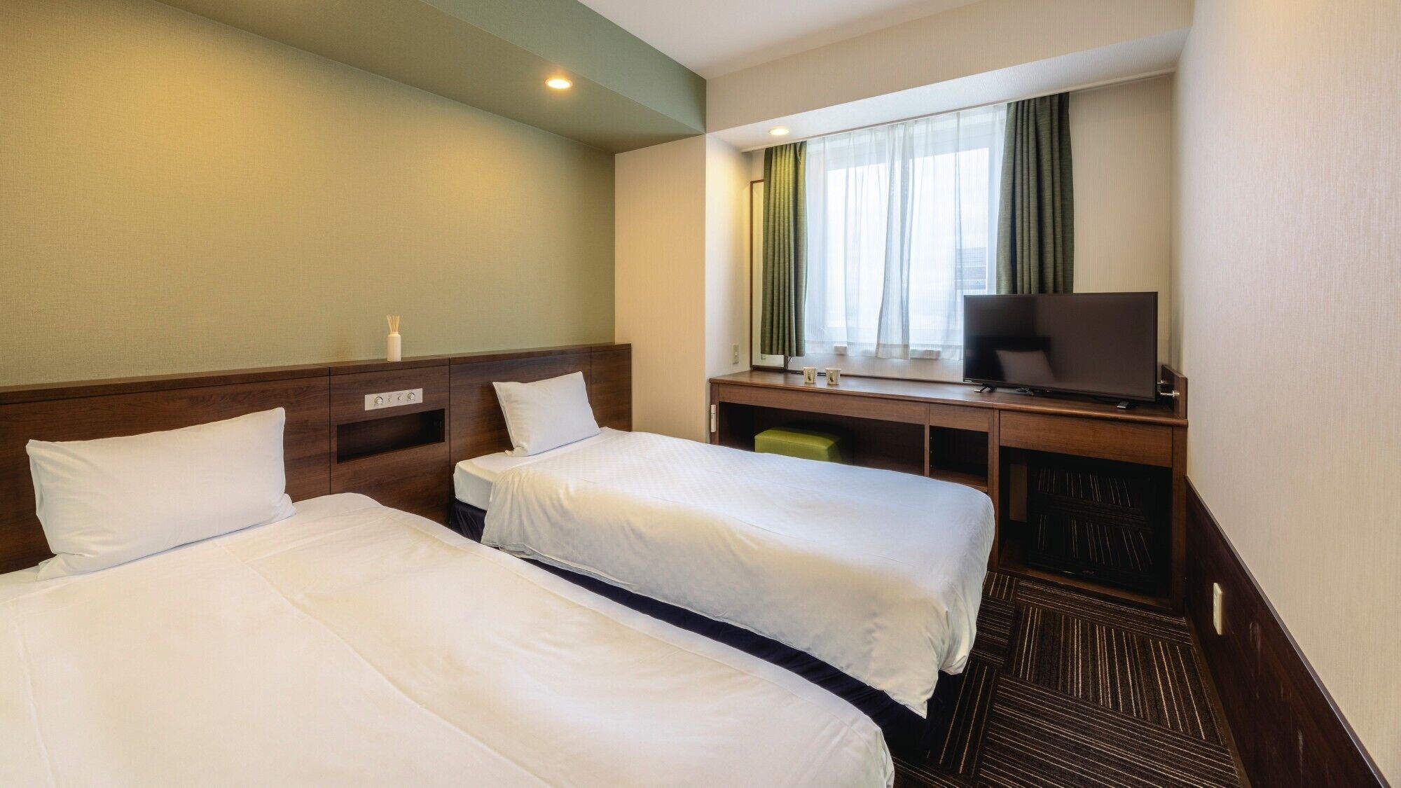 A hotel room with two beds, a desk with a TV, and a window with curtains.