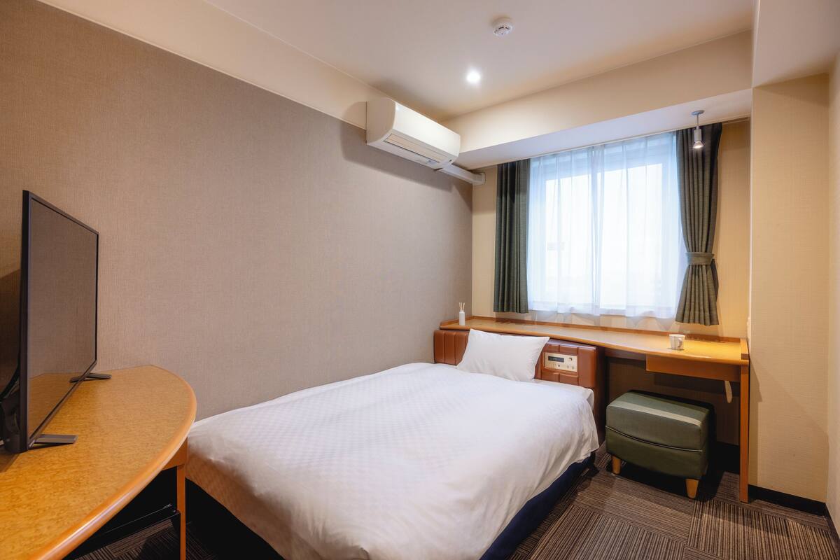 Une chambre d’hôtel comprenant un lit, un bureau, une chaise, une petite table, un appareil de climatisation, une fenêtre avec des rideaux et un téléviseur.