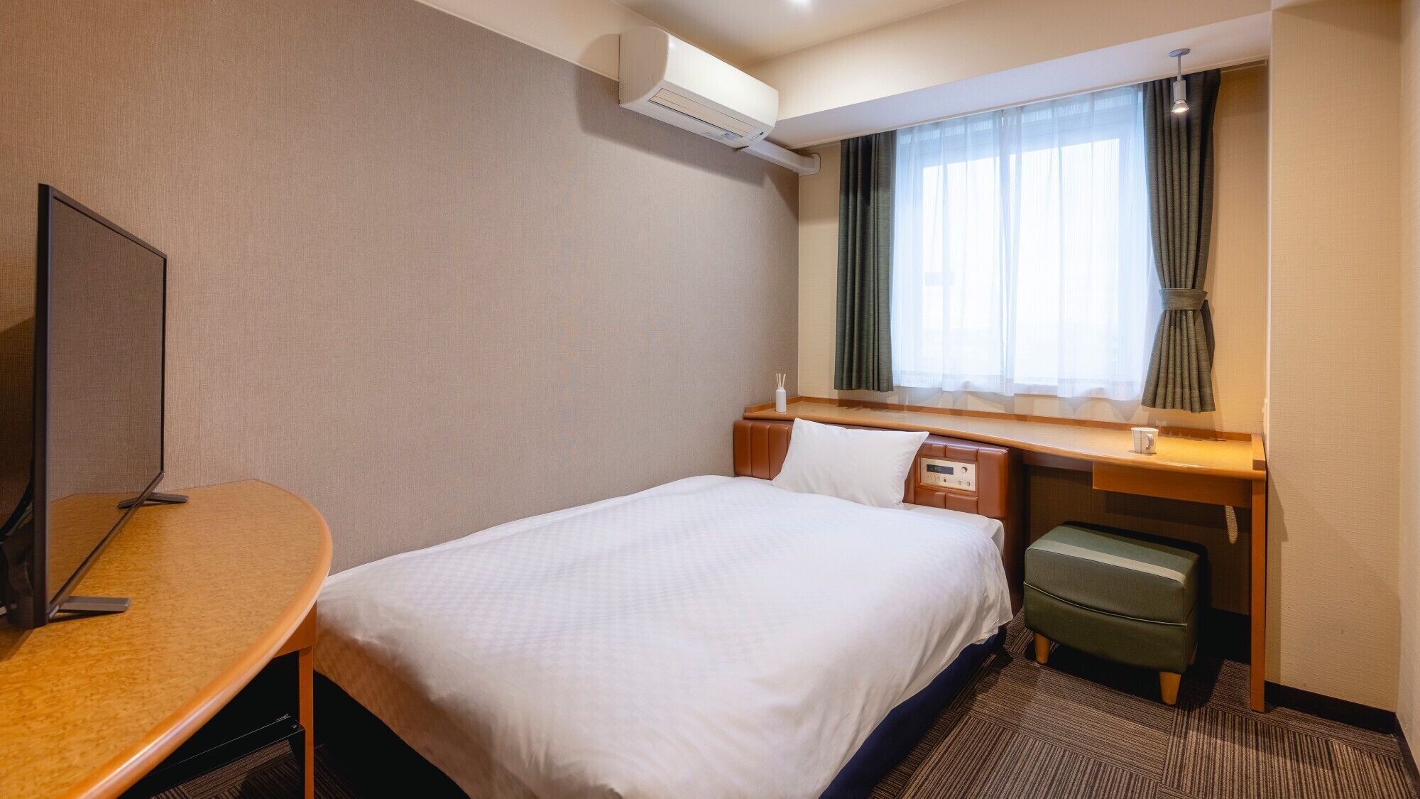 A hotel room with a bed, a desk, a chair, a TV, a window with curtains, and an air conditioning unit.
