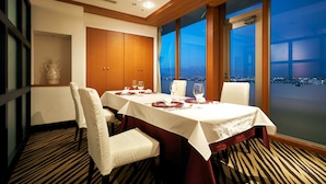A dining area with a table set for four, large windows offering a city view, and a vase on a shelf.