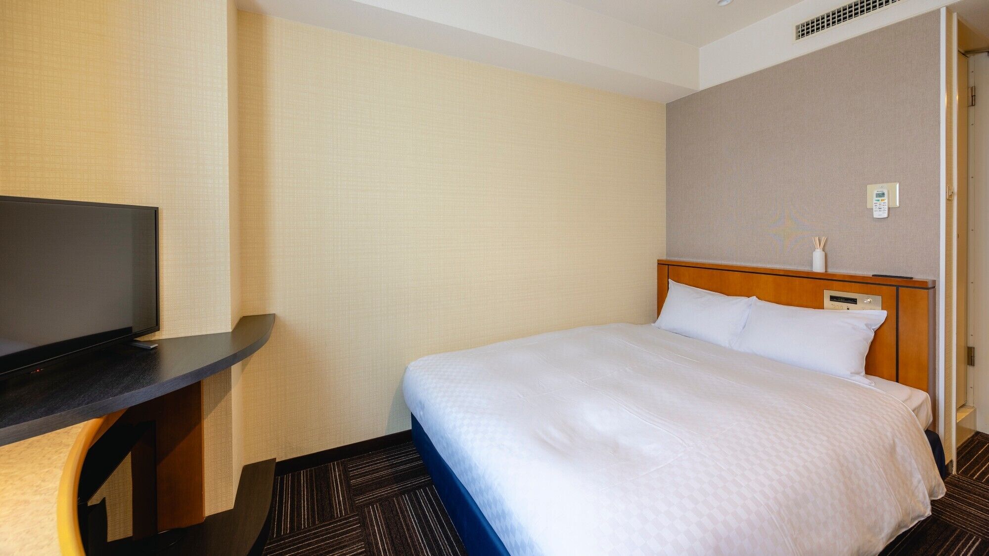 A hotel room with a bed, a TV on a stand, a bedside table, and a wall-mounted thermostat.