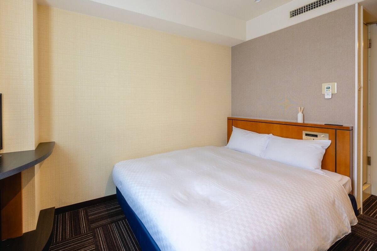 Une chambre d’hôtel avec un lit, un téléviseur posé sur un meuble, une table de chevet et un thermostat fixé au mur.