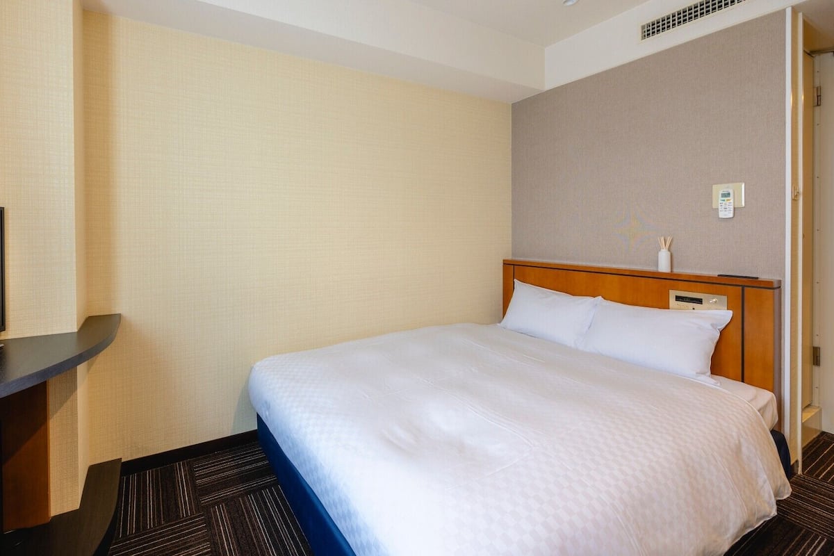 A hotel room with a bed, a TV on a stand, a bedside table, and a wall-mounted thermostat.