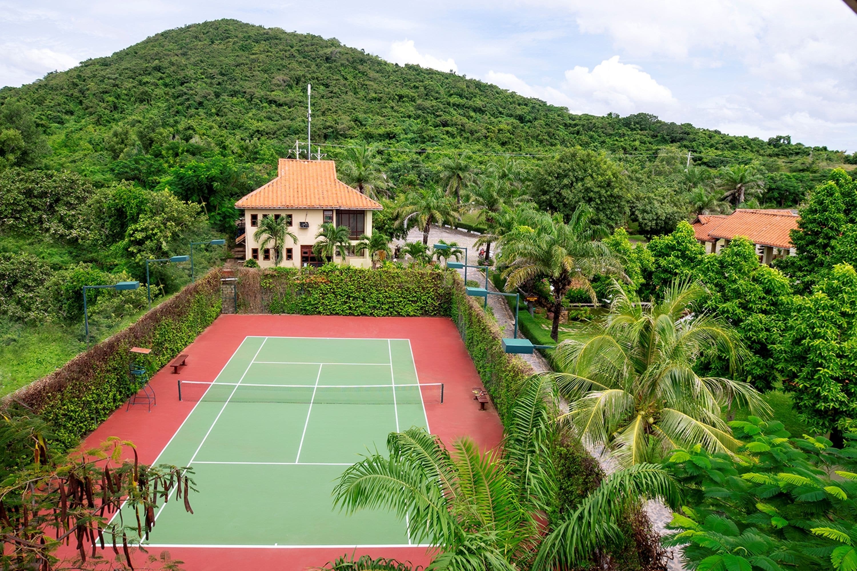 Una cancha de tenis rodeada de abundante vegetación y una colina al fondo.