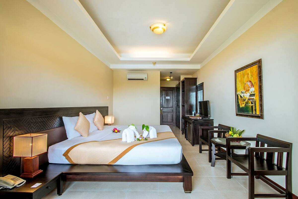 A hotel room with a large bed, bedside table with a lamp, a desk with a chair, a television, and a painting on the wall.