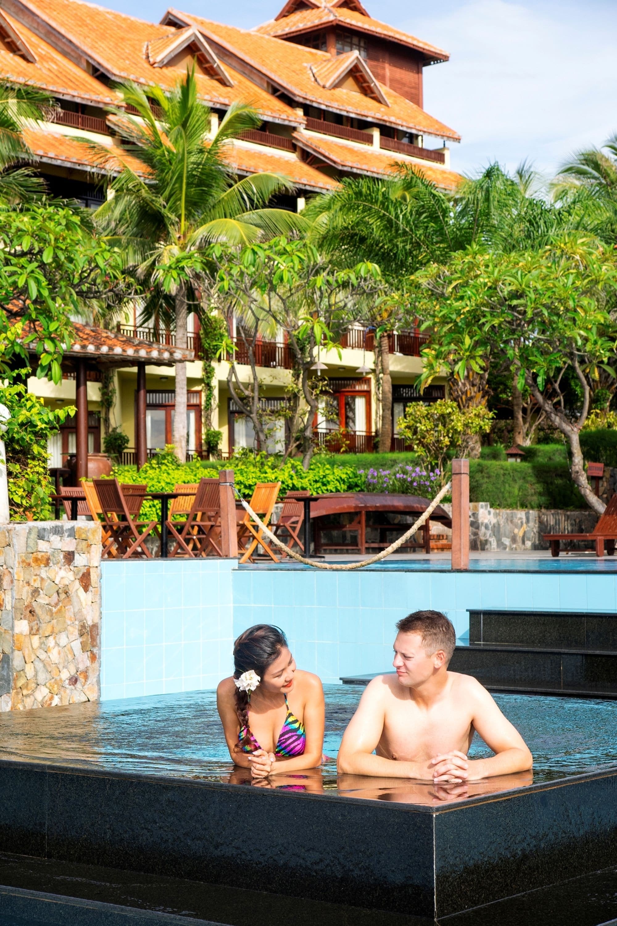 Area piscina con una coppia, l'edificio di un resort tropicale e vegetazione rigogliosa.