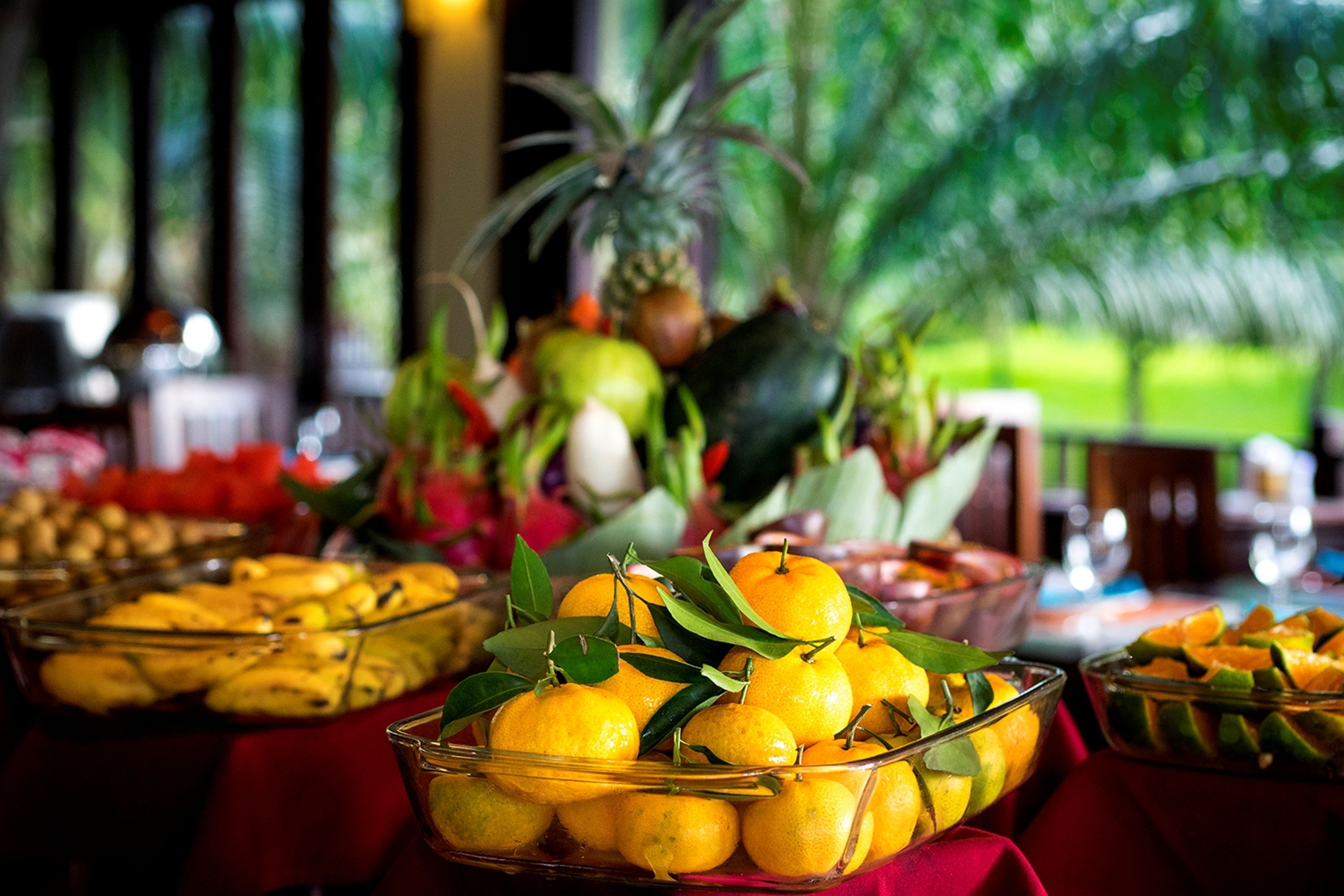 A buffet table with various fruits and dishes, including oranges and a pineapple, with a view of greenery in the background.