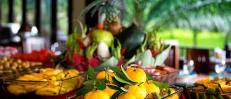 A buffet table with various fruits and dishes, including oranges and a pineapple, with a view of greenery in the background.
