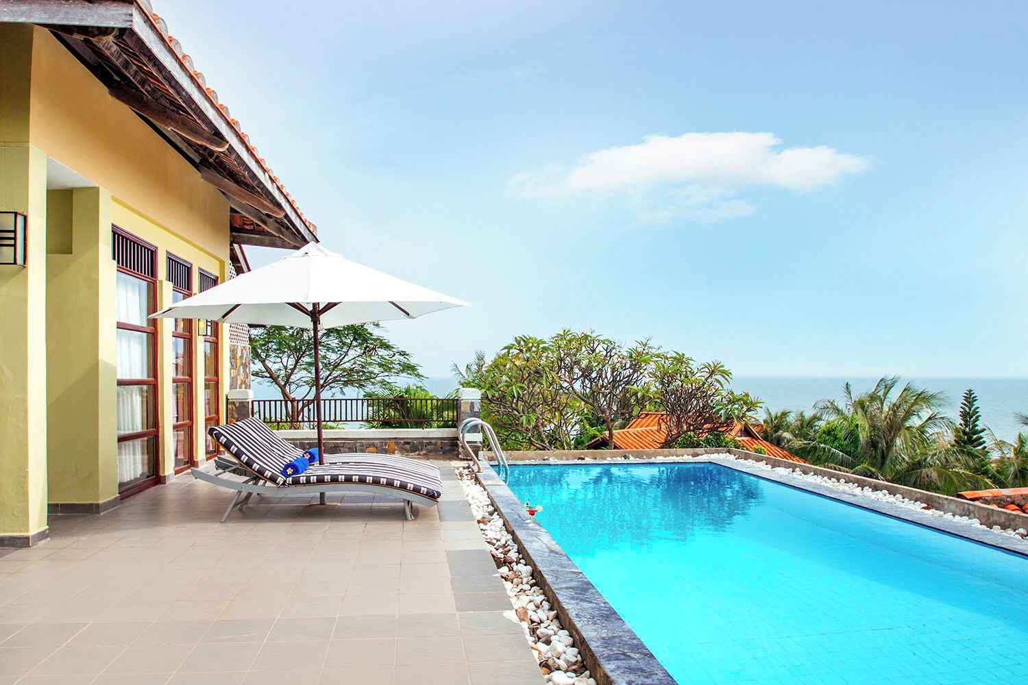 A swimming pool with a view of the ocean, a lounge area with a sun umbrella, and a building with large windows.