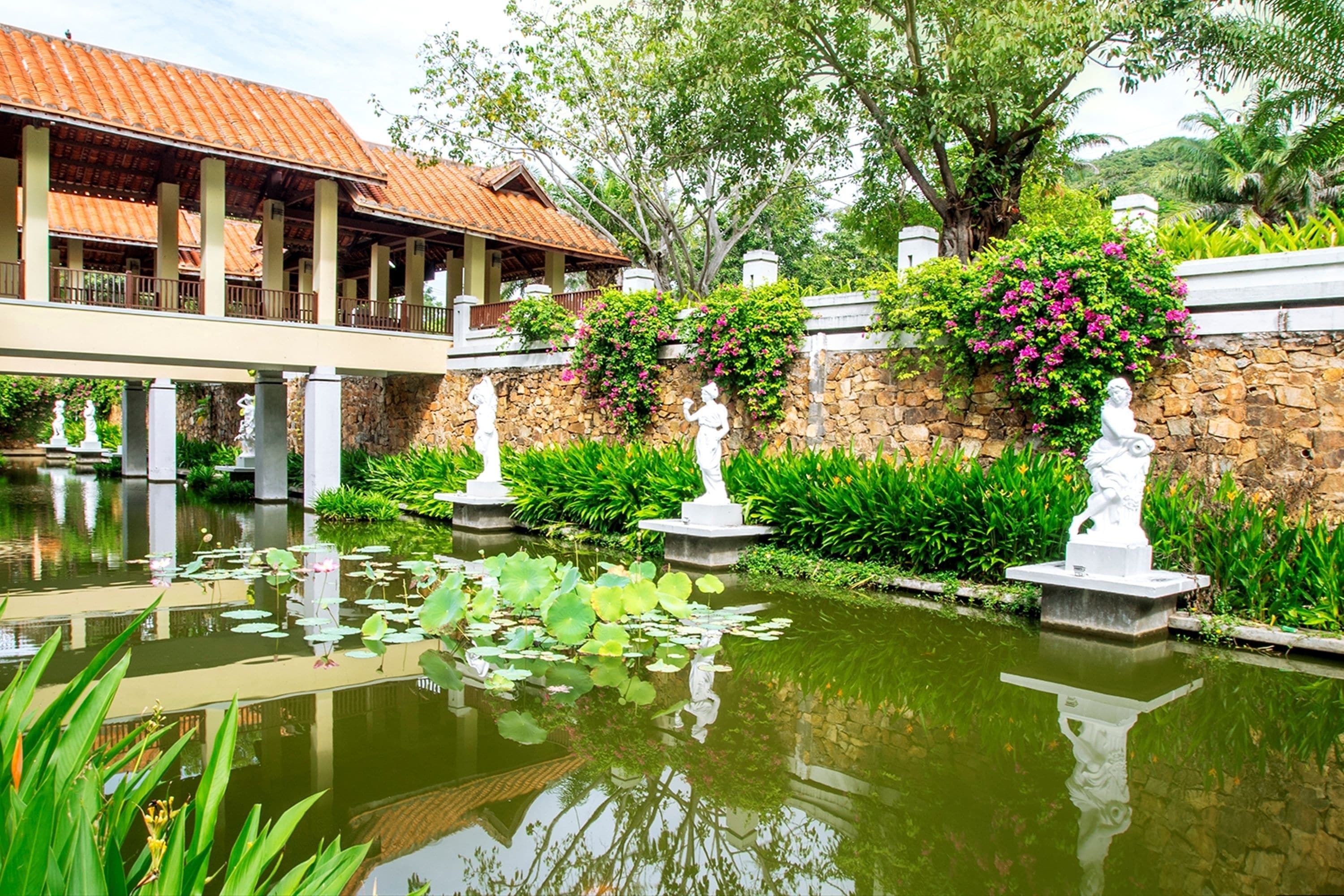 A serene garden with a pond, stone statues, and a traditional building with a tiled roof.