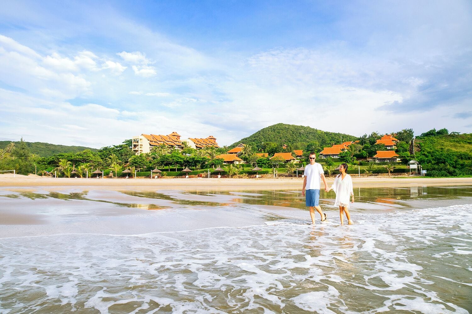 A beach with a resort in the background, featuring multiple buildings with red roofs and lush greenery.