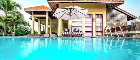 A poolside area with lounge chairs, a covered seating area, and a building with a tiled roof.