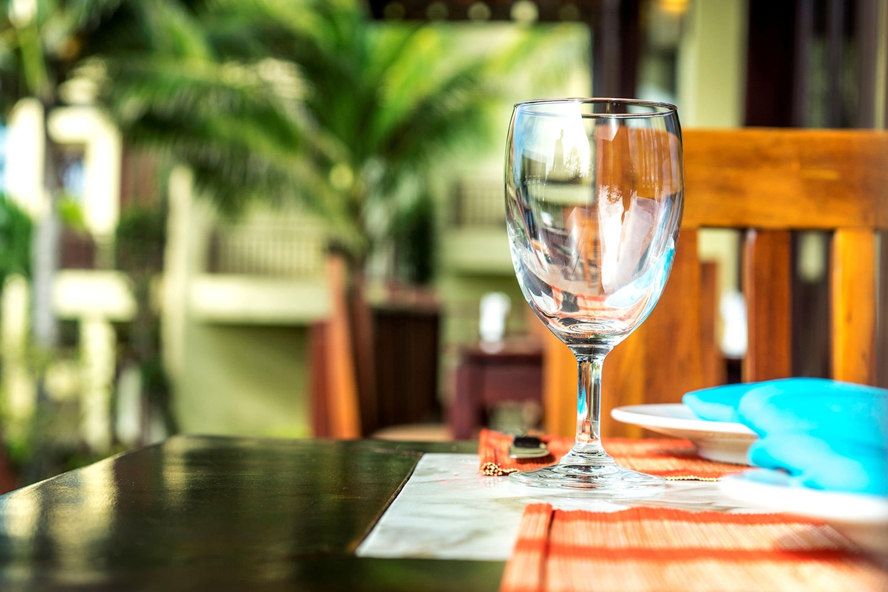 A glass of water on a table with a placemat, a wooden chair, and a blue plate in the background.