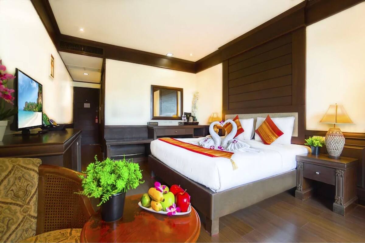 Hotelkamer met een groot bed, nachtkastje met een lamp, een klein tafeltje met fruit en een plant, en een televisie op een dressoir.