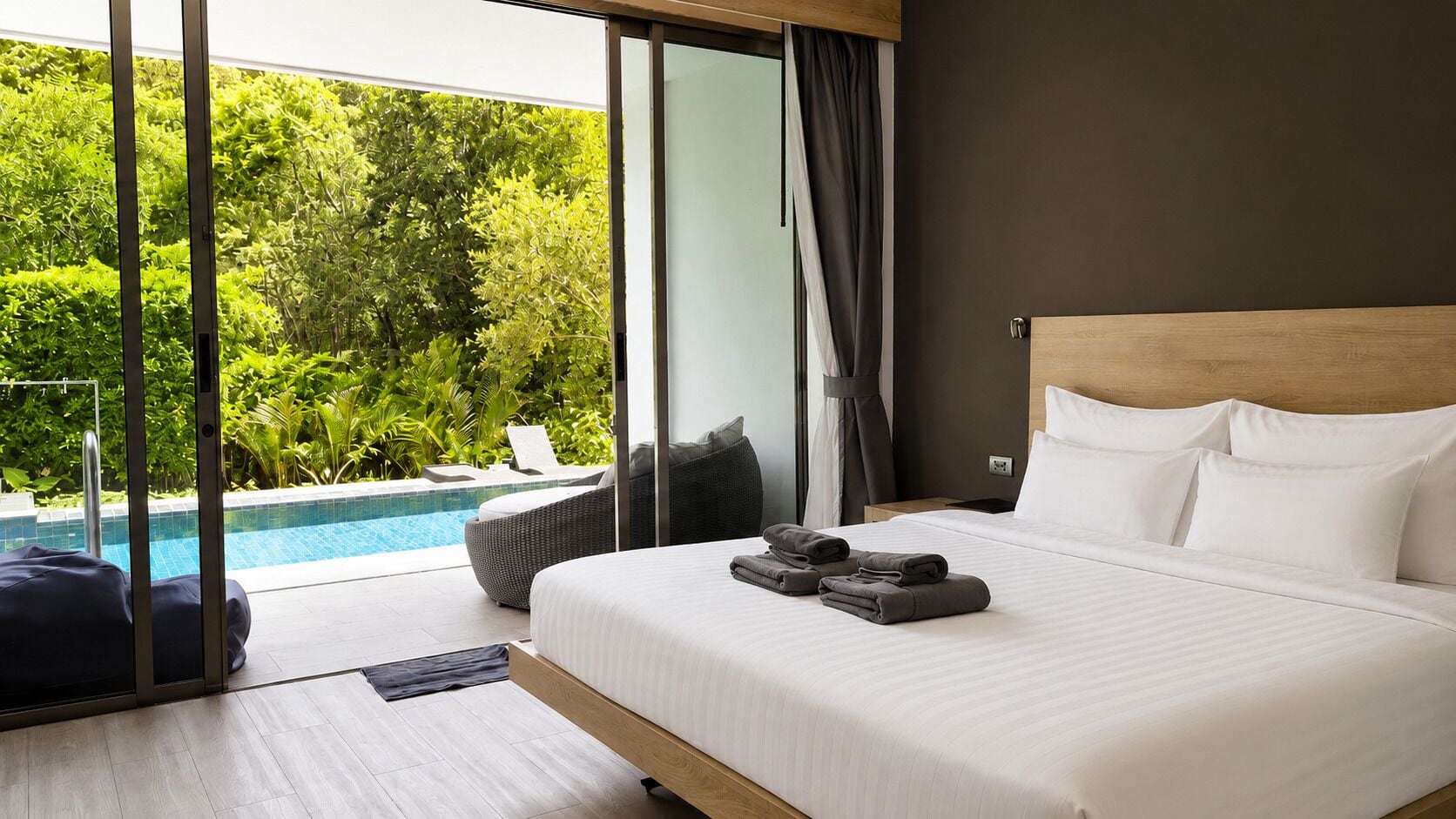 A modern hotel room with a large bed, a view of a swimming pool, and a balcony with a view of lush greenery.