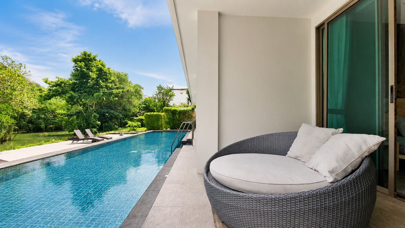 A poolside area with a wicker chair and lounge chairs, a clear blue pool, and a view of greenery and a building in the background.