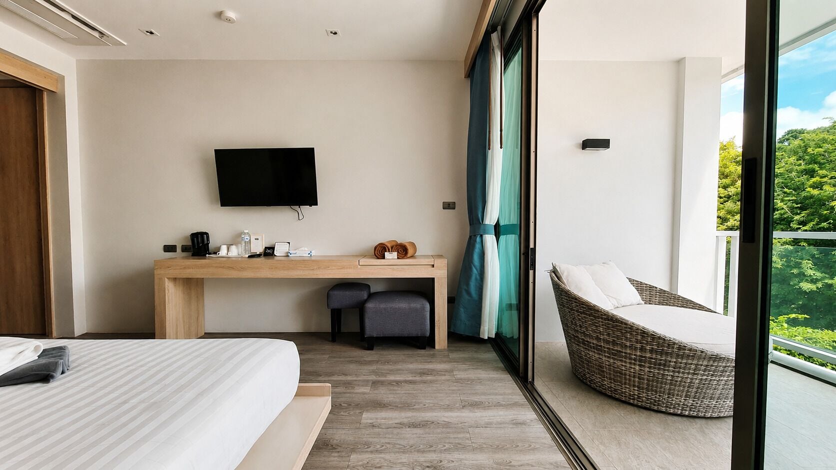 A hotel room with a bed, a desk with a flat-screen TV, a wicker chair, and a balcony with a view.