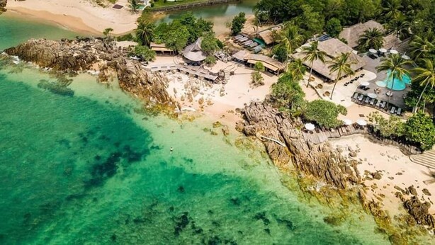 Vista aérea de un complejo turístico en la playa con aguas turquesas cristalinas, cabañas de techo de paja, palmeras y una zona de piscina.