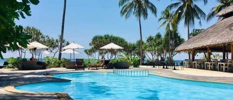 A pool area with lounge chairs and umbrellas, surrounded by palm trees and a thatched-roof structure.