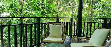 A covered outdoor seating area with wicker chairs and cushions, surrounded by a black railing, overlooking a lush green forest.