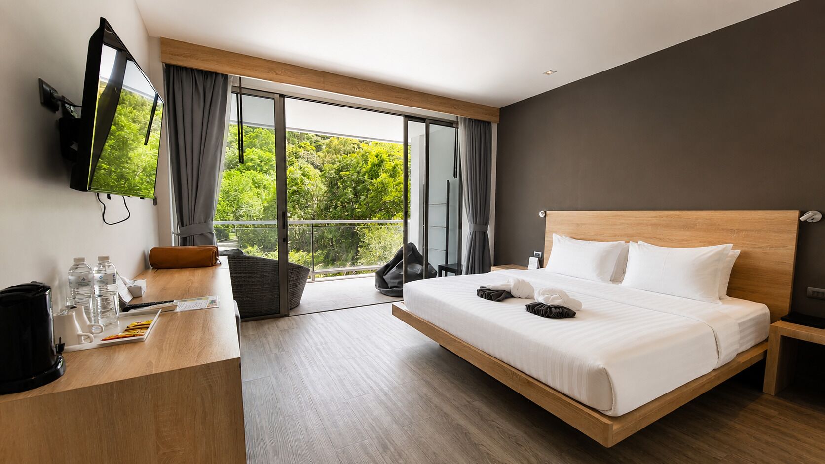 A modern hotel room with a large bed, a desk with a coffee maker, a flat-screen TV, and a balcony with a view of greenery.