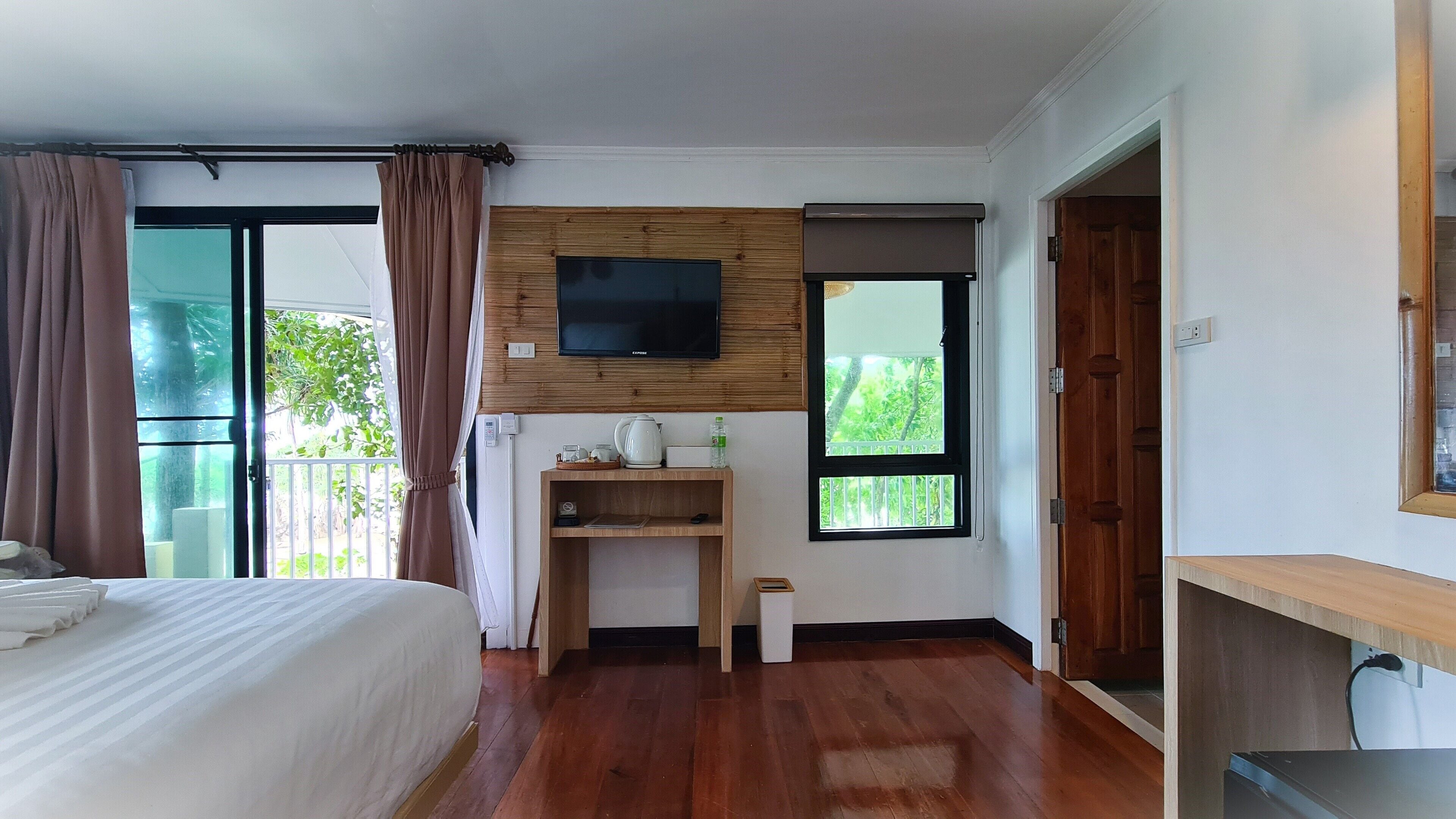 A hotel room with a bed, a TV mounted on a wooden panel, a small wooden shelf with a kettle and a bottle, a window with a view of greenery, and a door leading to another room.