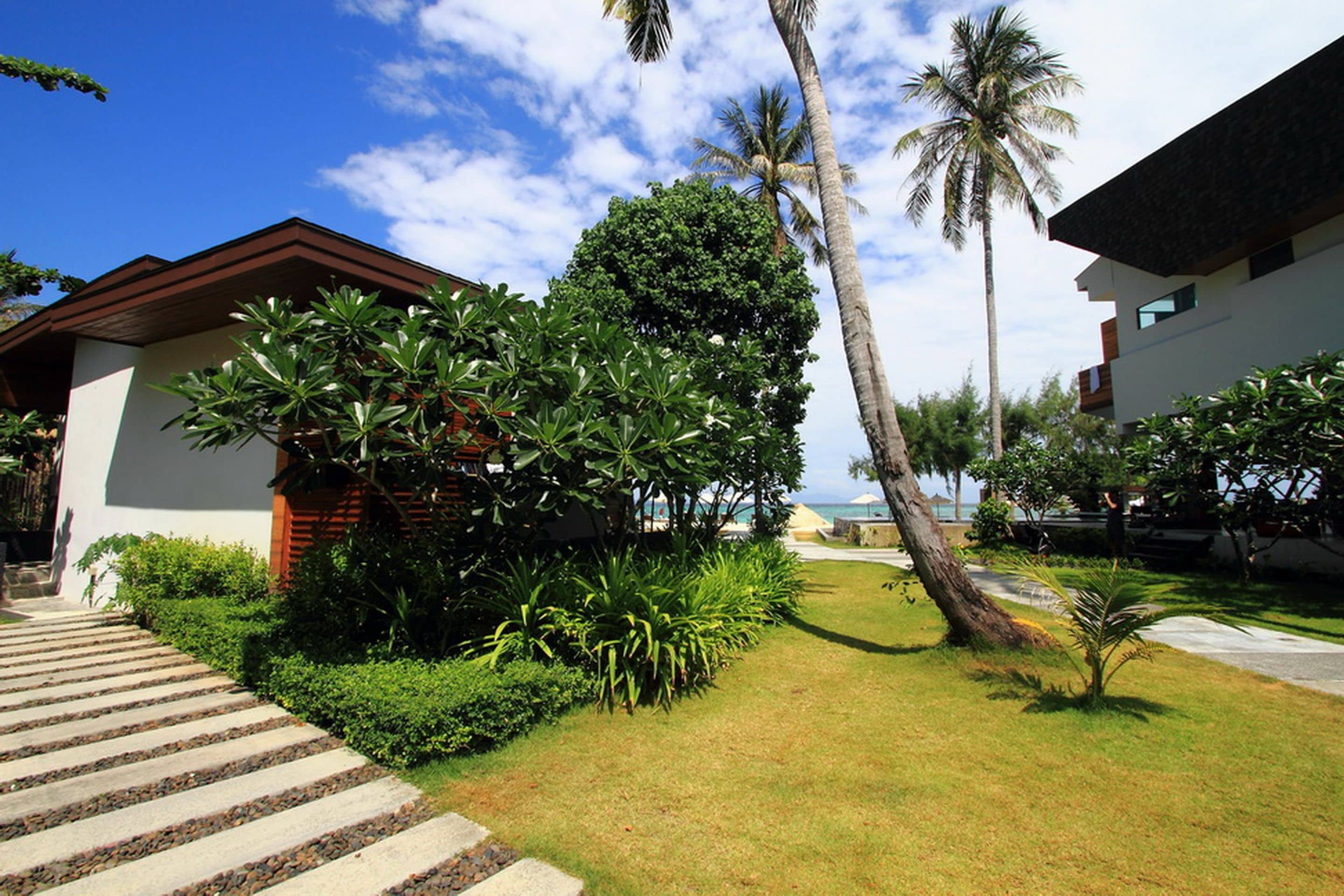A modern building with a stone pathway leading to the entrance, surrounded by lush greenery and palm trees.