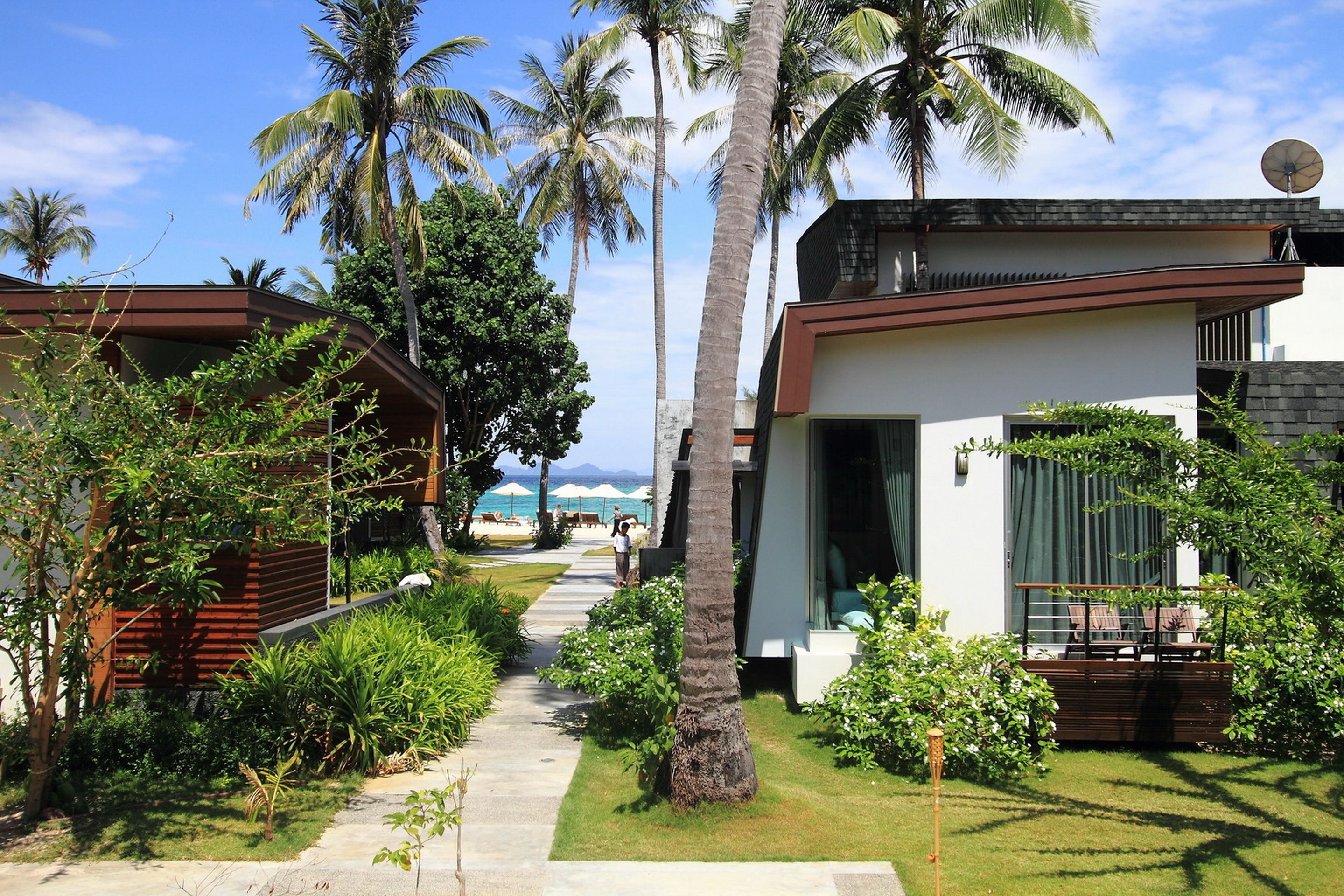 A pathway leading to a modern building with a balcony, surrounded by lush greenery and palm trees.