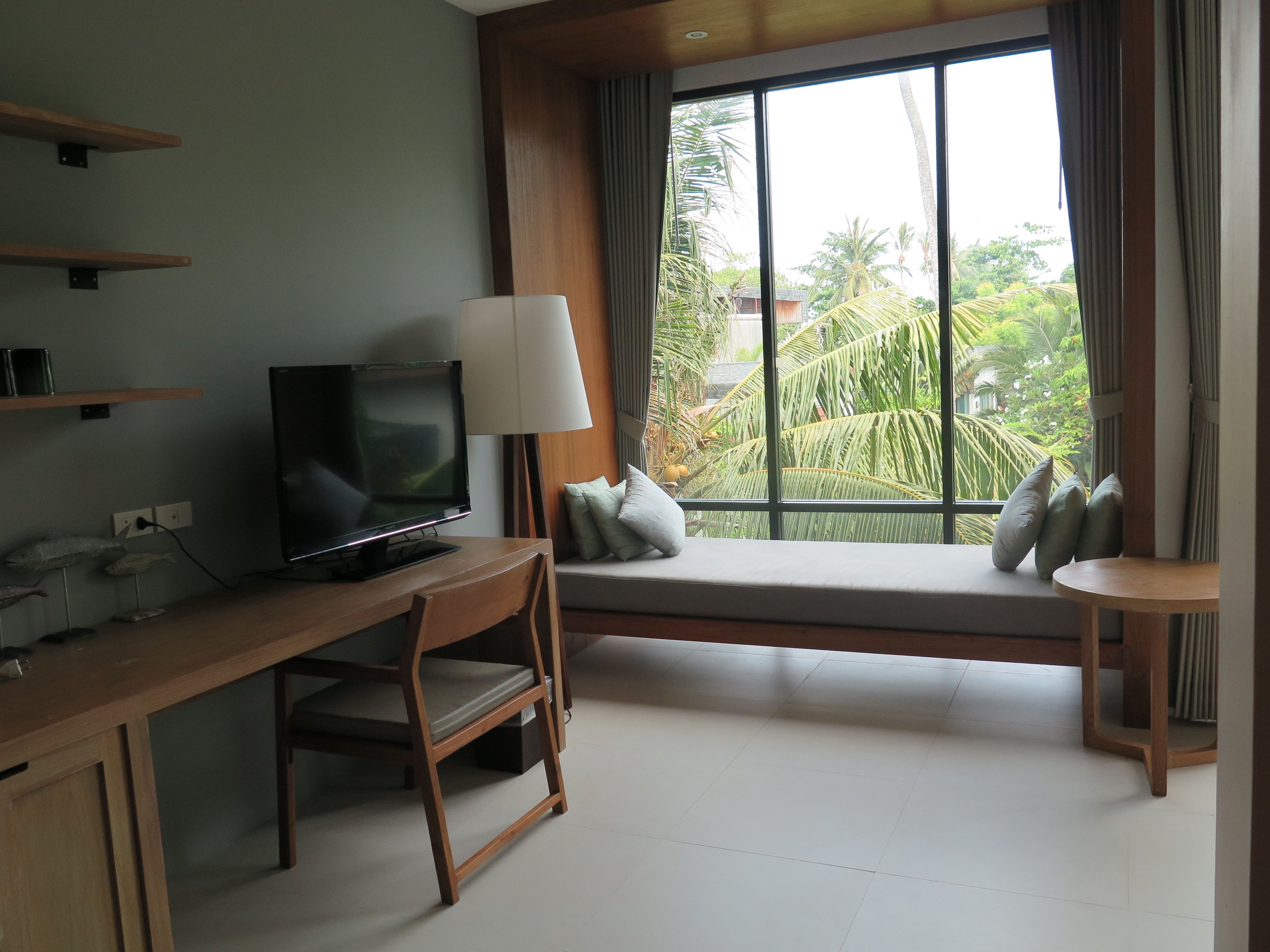 A modern hotel room with a wooden desk, a television, a large window with a view of palm trees, and a small seating area with cushions.