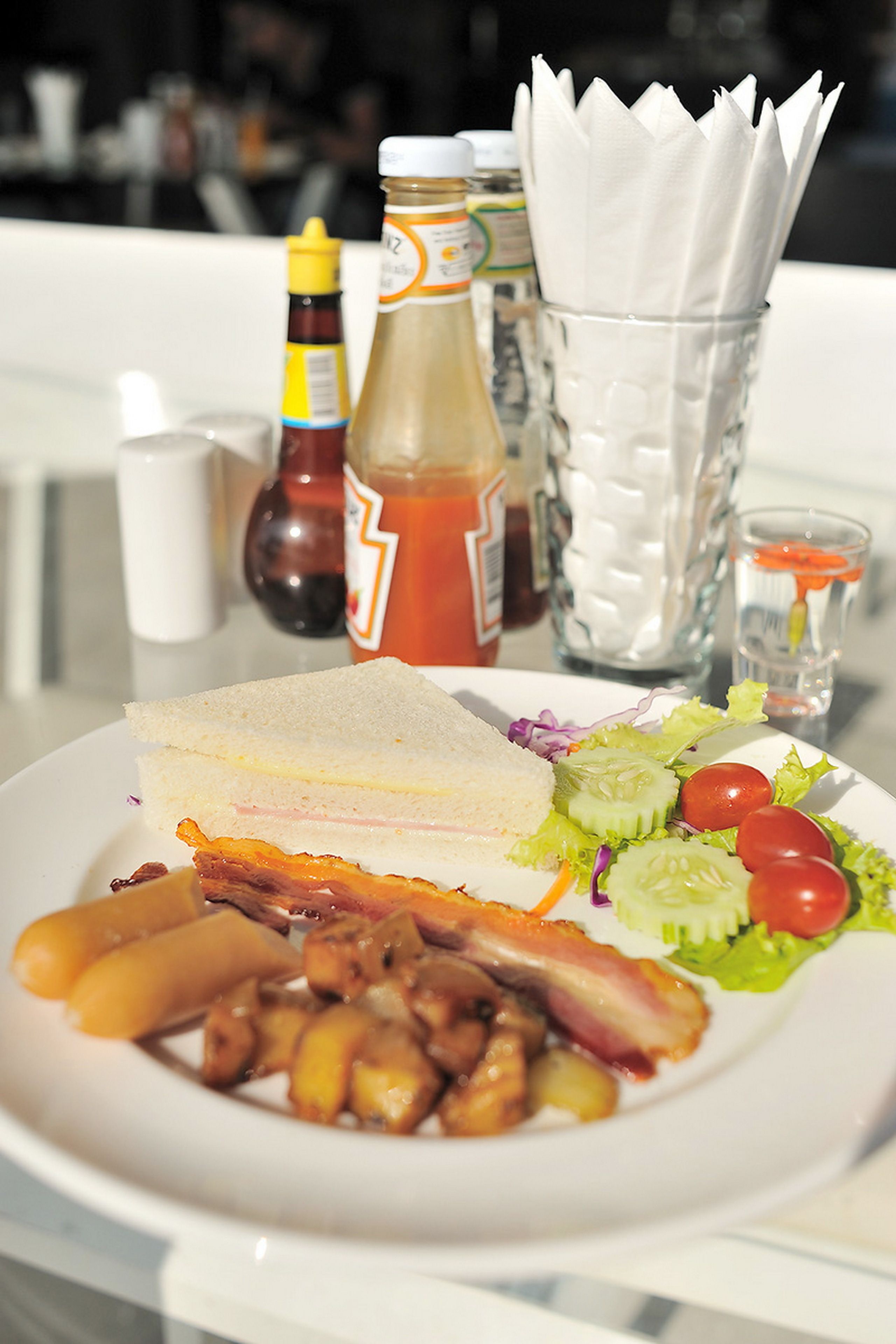 Un plato de comida con un sándwich, tocino, salchichas y una ensalada, acompañado de una servilleta y salsas sobre la mesa.