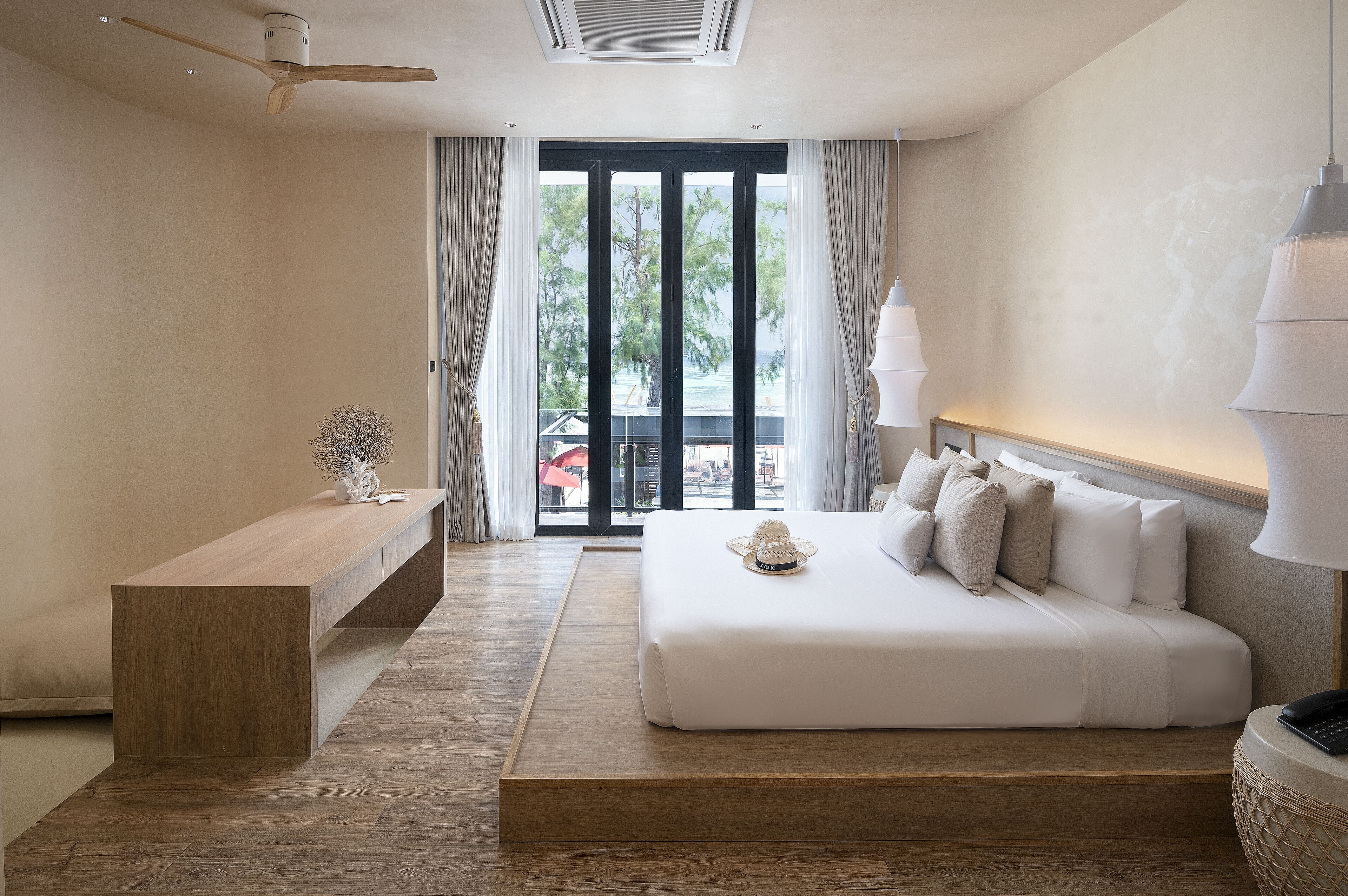 A modern hotel room with a large bed, a wooden bench, and a view of the outdoors through a sliding door.