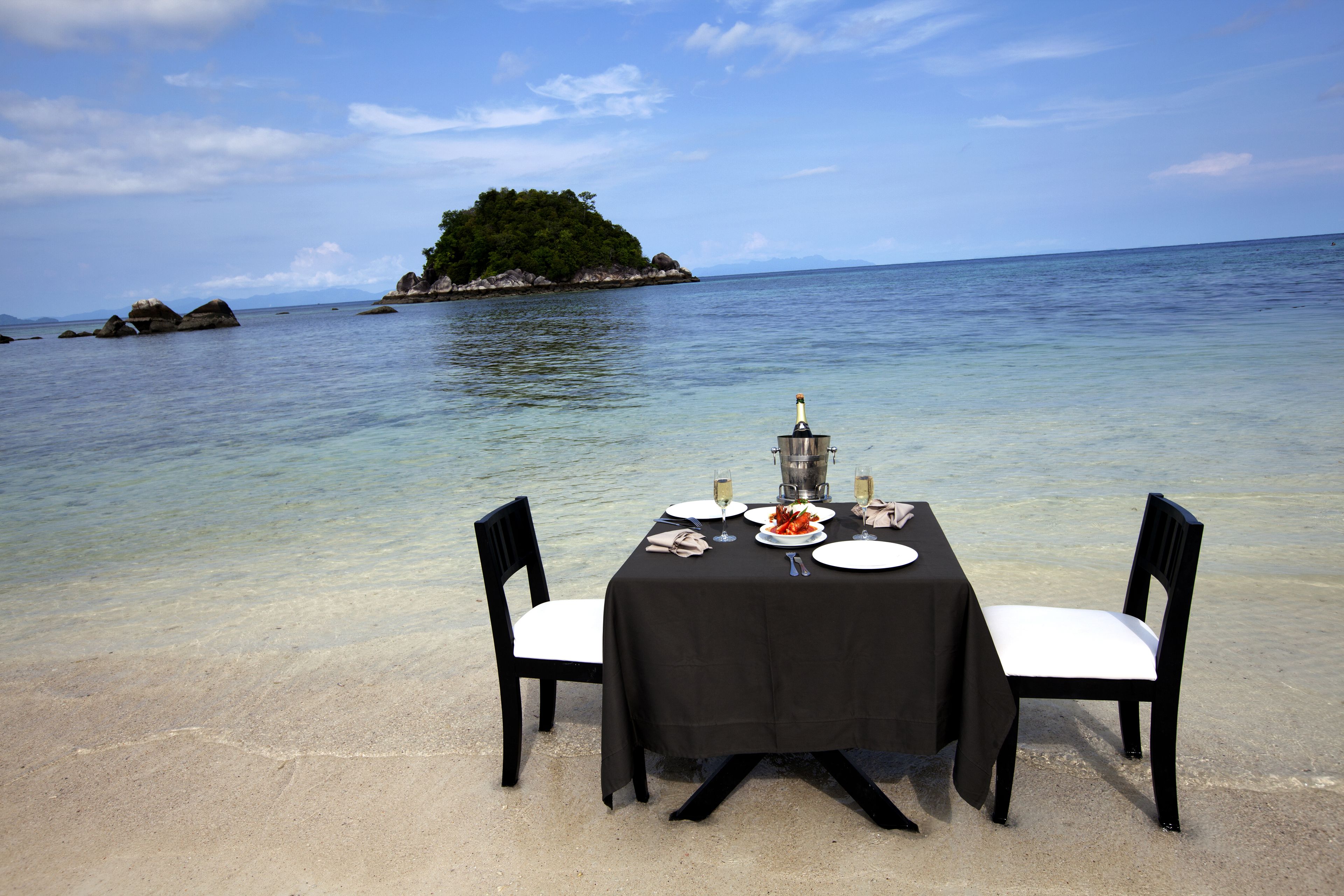 A table set for two with a bottle of champagne, plates, and cutlery on a beach with clear water and a distant island.