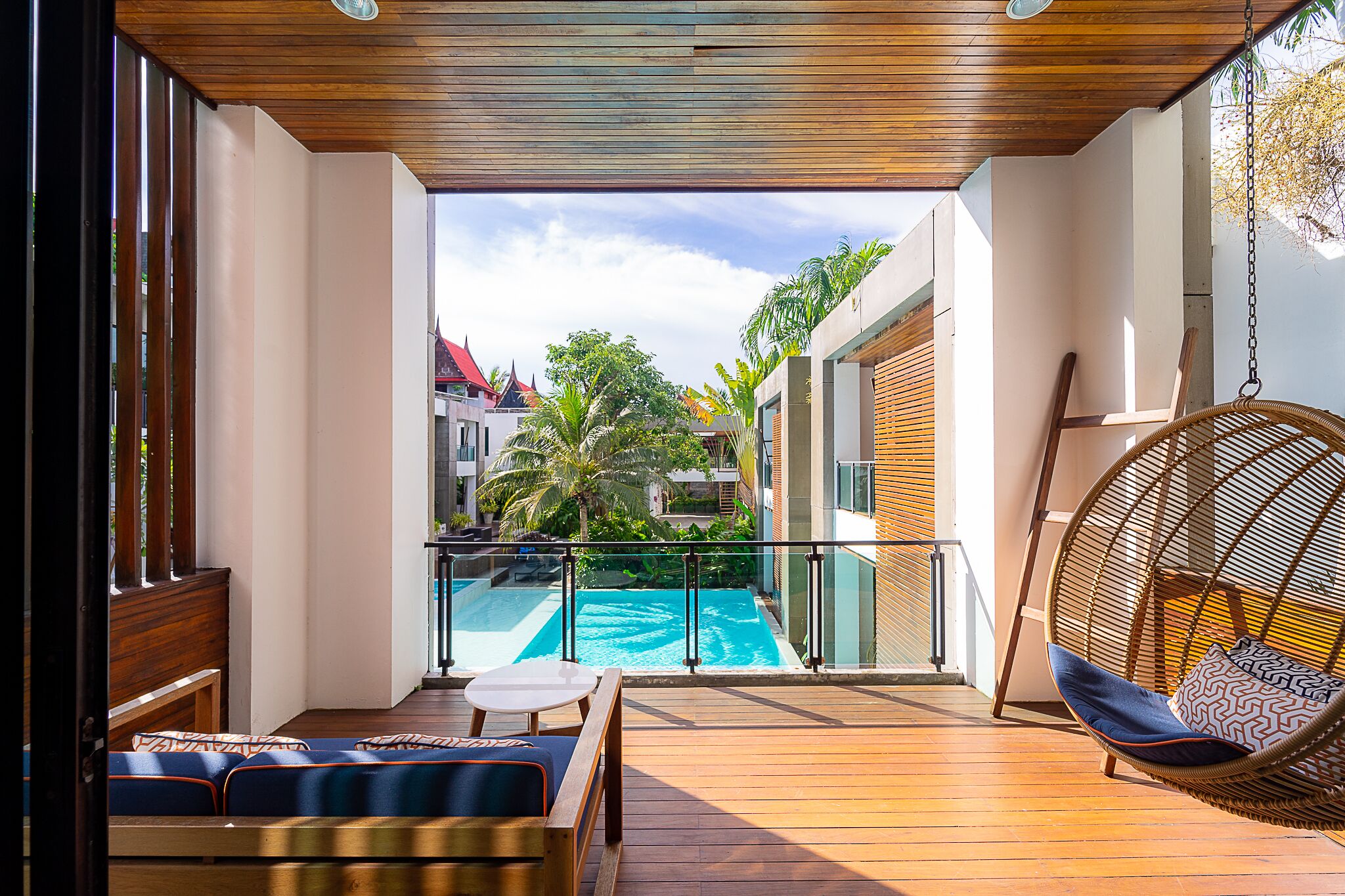 A balcony with a wooden floor, a hanging chair, a small table, and a view of a swimming pool and buildings.