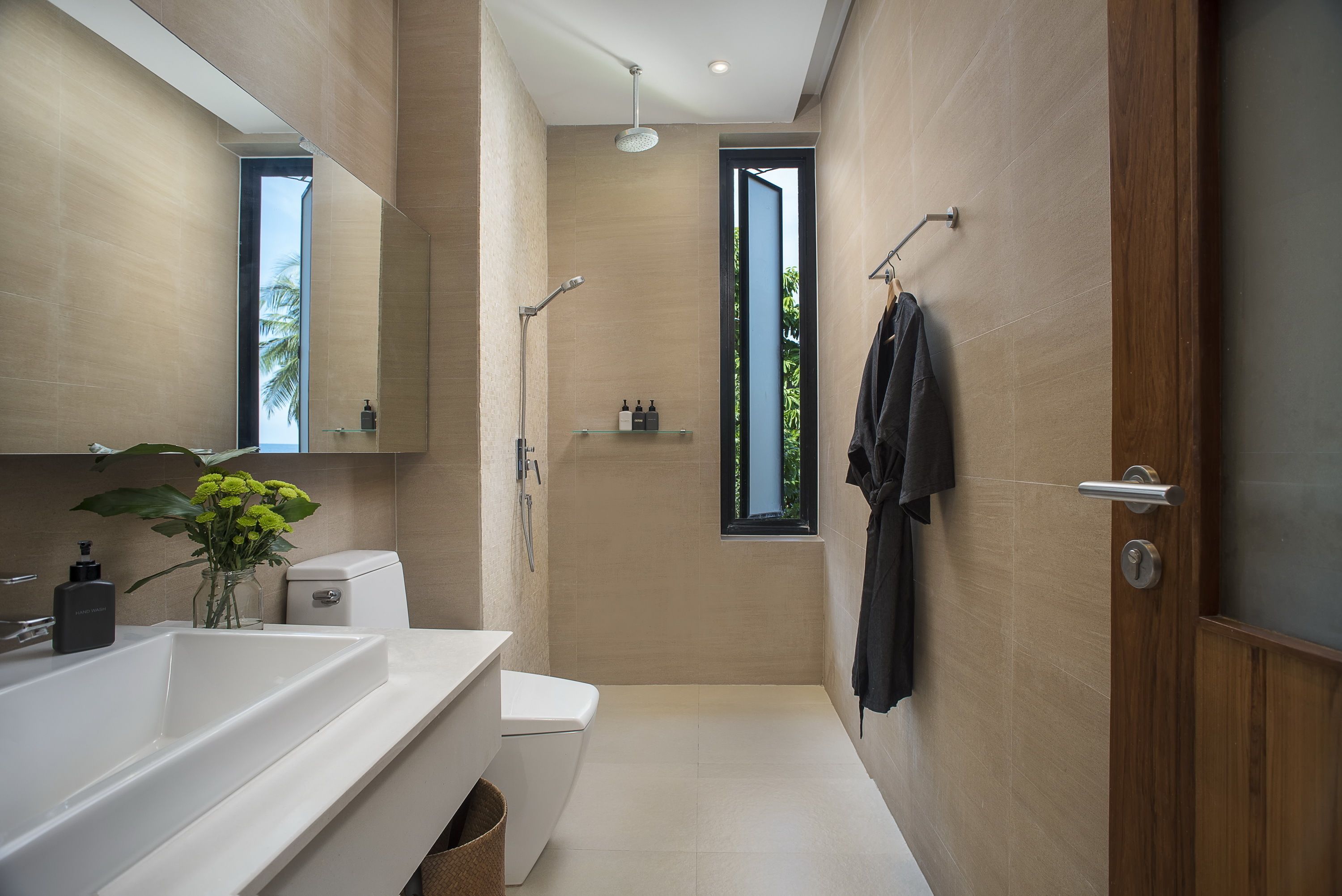 A modern bathroom with a large sink, toilet, and shower. There is a window providing natural light and a view of greenery outside.