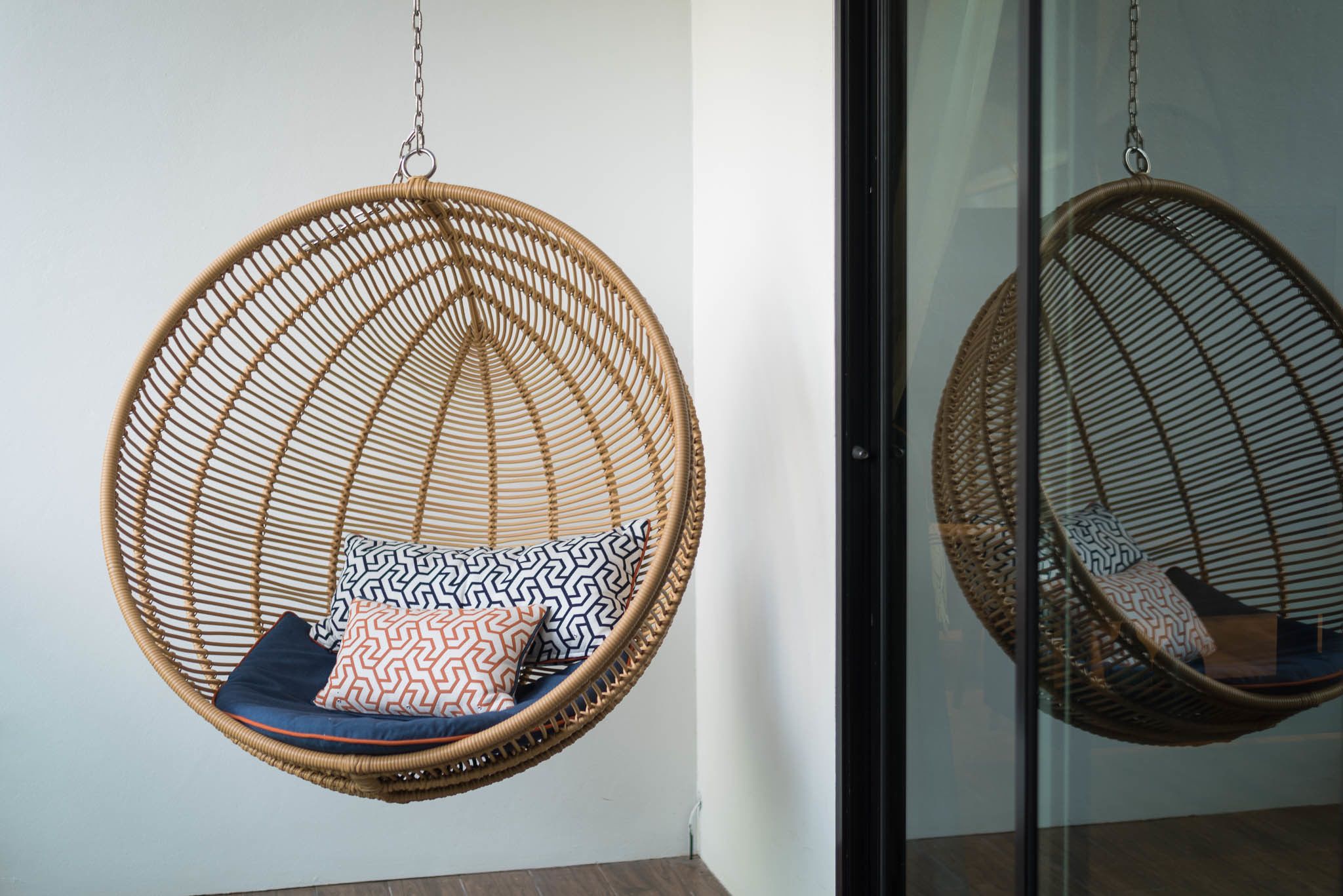 Two wicker hanging chairs with patterned cushions against a white wall, one with a glass door reflection.