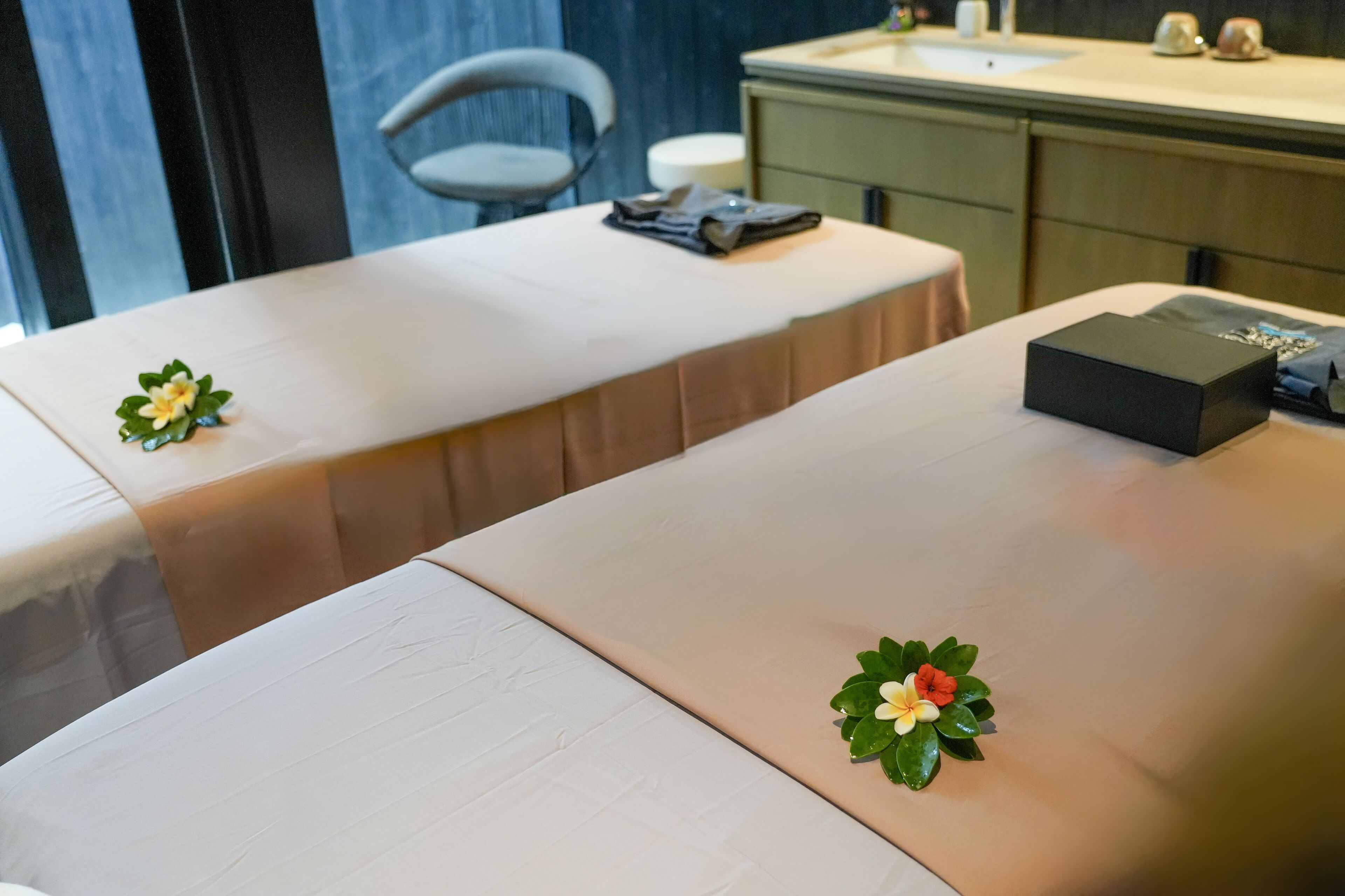 Zwei Massagetische mit beiger Auflage, eine kleine Pflanze und eine schwarze Box auf einem Tisch.