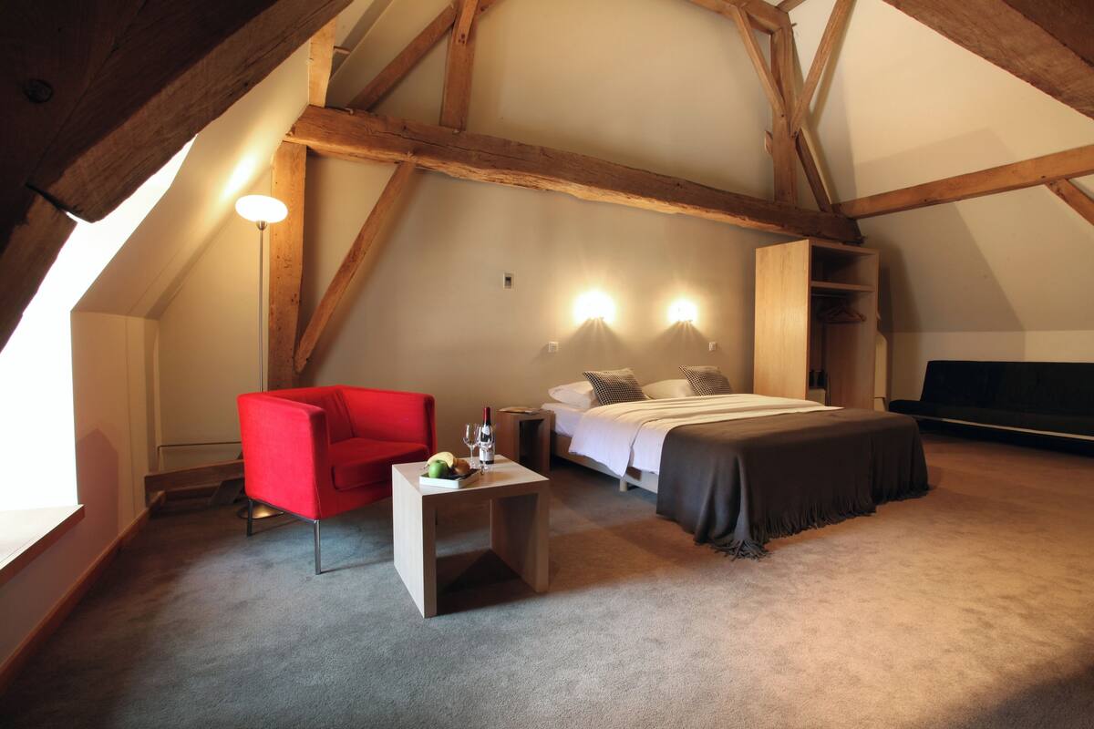 A bedroom with a sloped ceiling, a bed with a dark bedspread, a red chair, a small table with a bottle and glasses, and a wall-mounted light fixture.