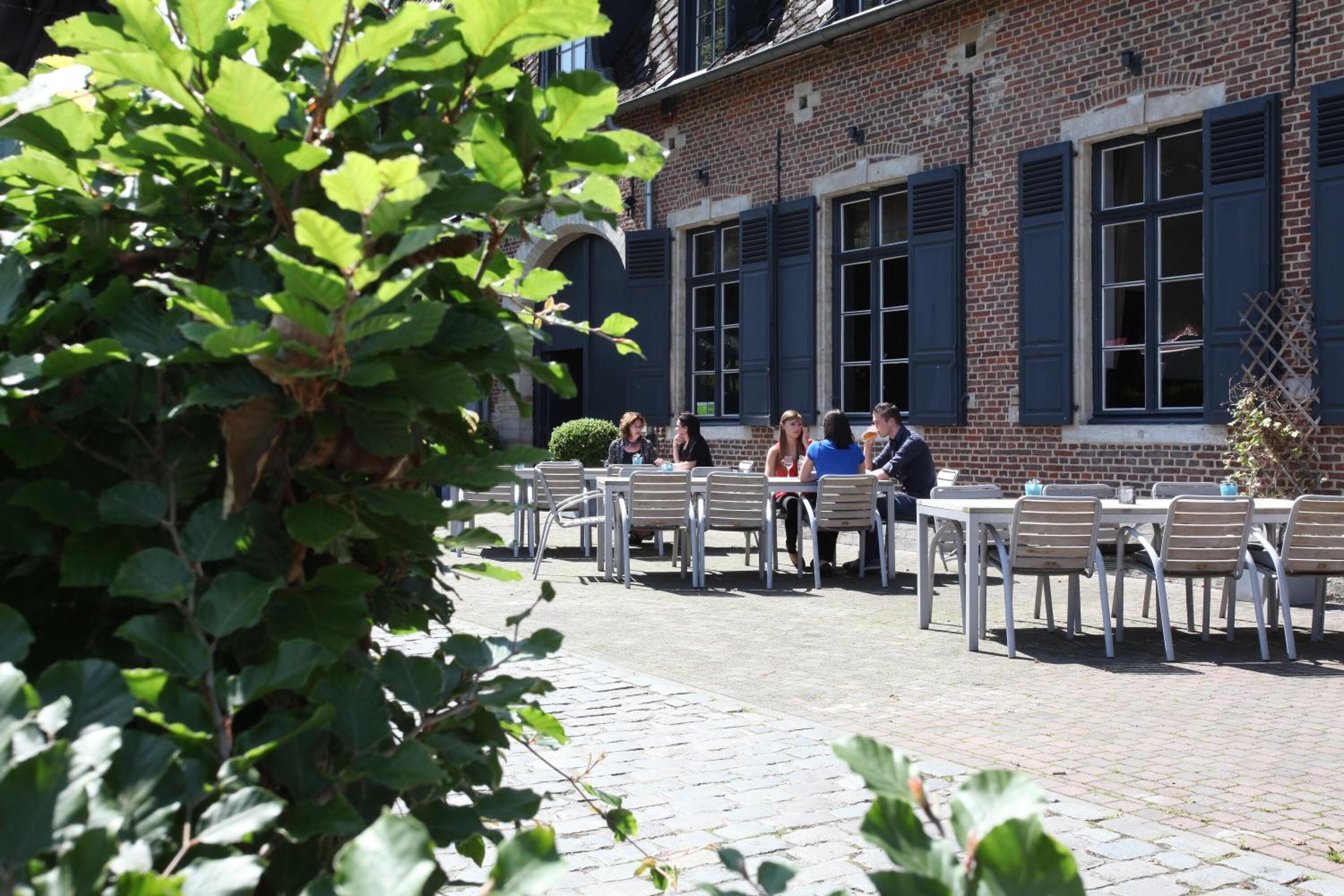 An outdoor cafe with tables and chairs, a brick building with shutters, and a tree in the foreground.