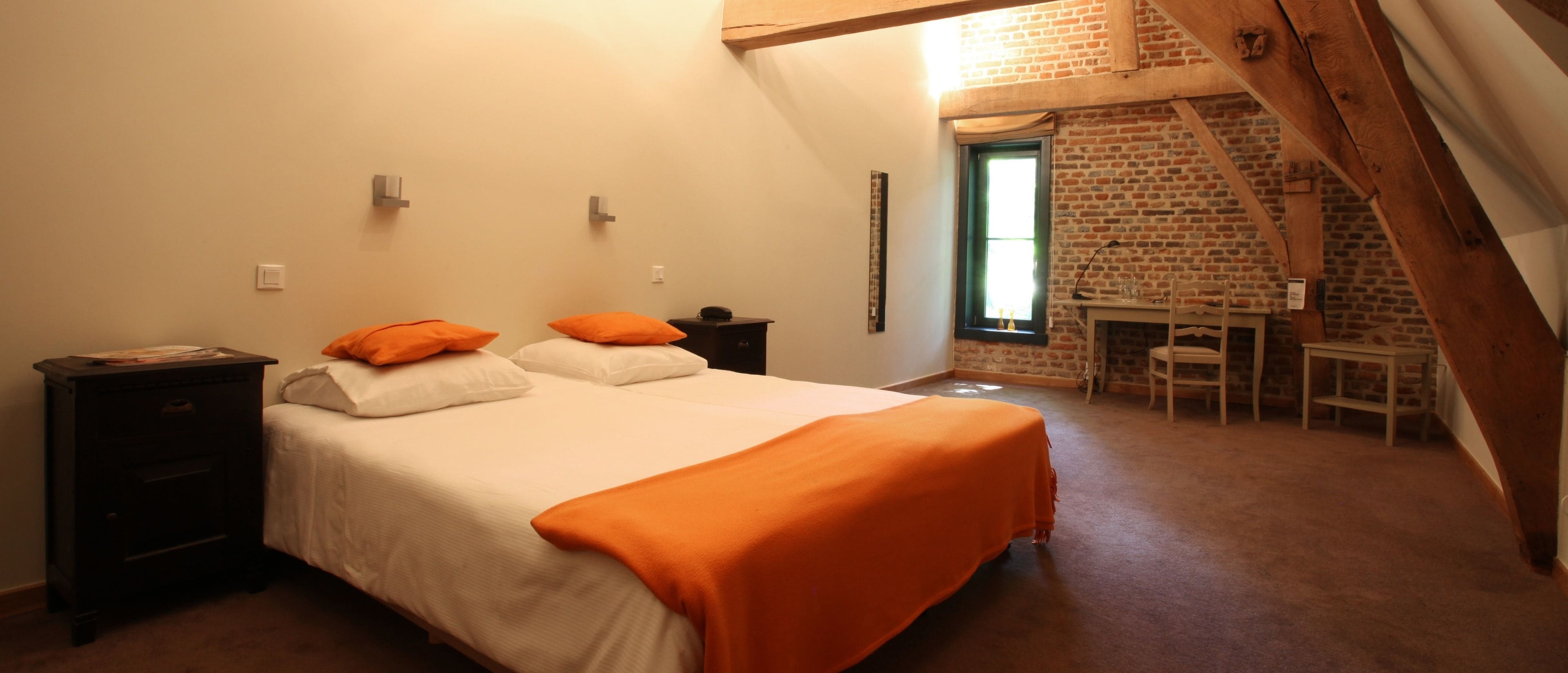 Un dormitorio con una cama, dos mesitas de noche, una manta naranja, un escritorio con una silla y una pared de ladrillo.
