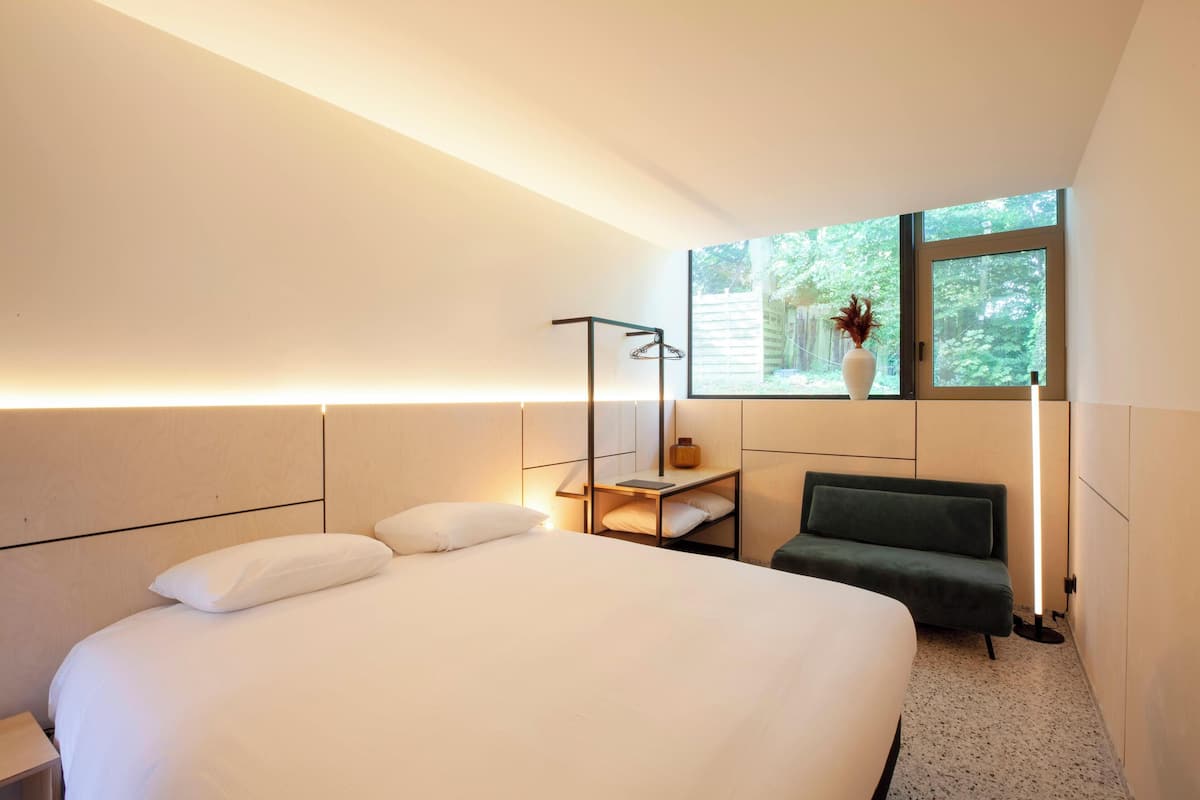 A modern hotel room with a large bed, a bedside table, a green sofa, a window with a view of greenery, and a vase with flowers on the table.