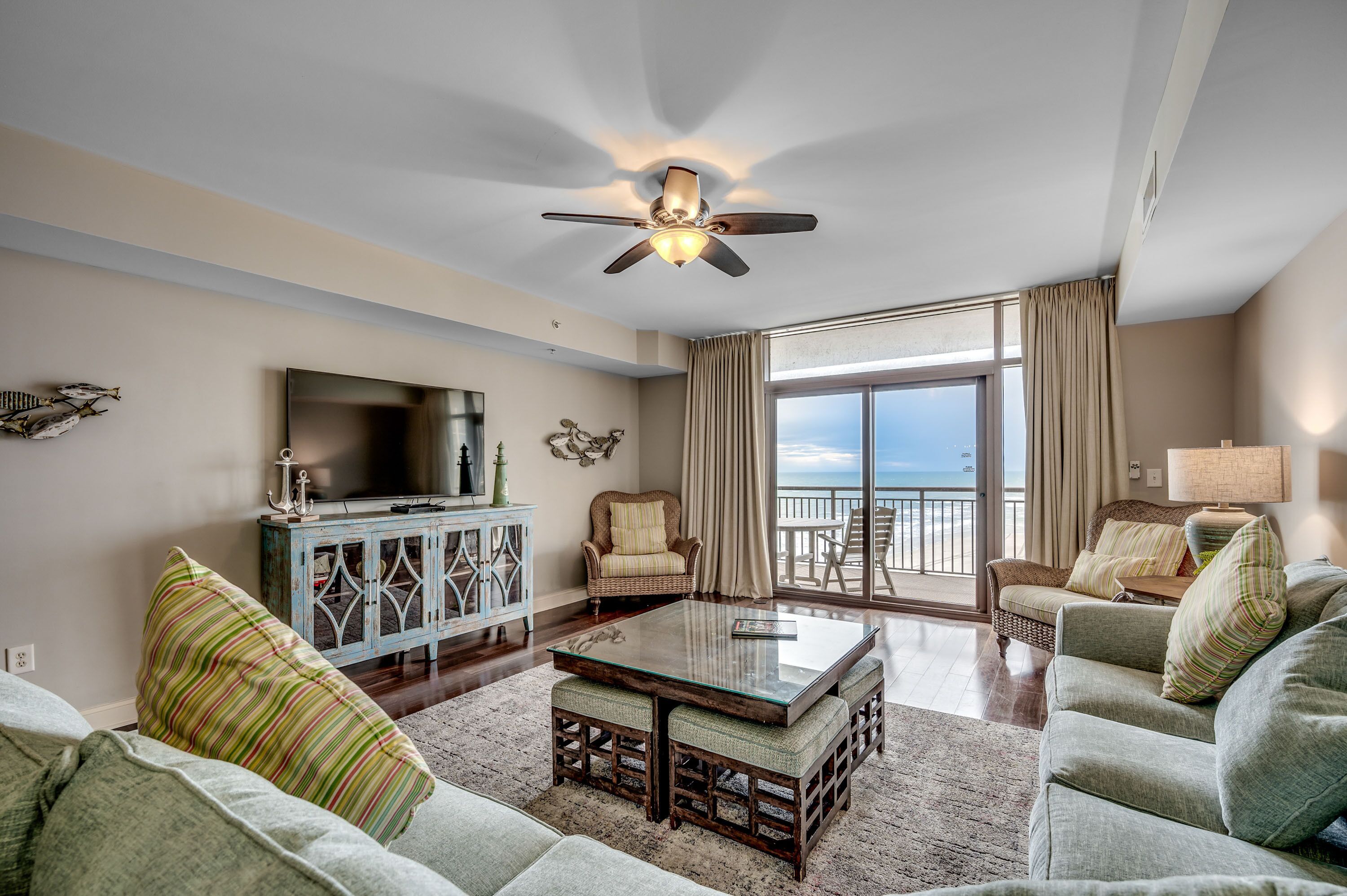 A spacious living room with a large flat-screen TV, a ceiling fan, and a balcony with a view of the ocean.