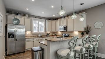 A modern kitchen with a stainless steel refrigerator, a microwave, and a sink. There are bar stools and a decorative light fixture.